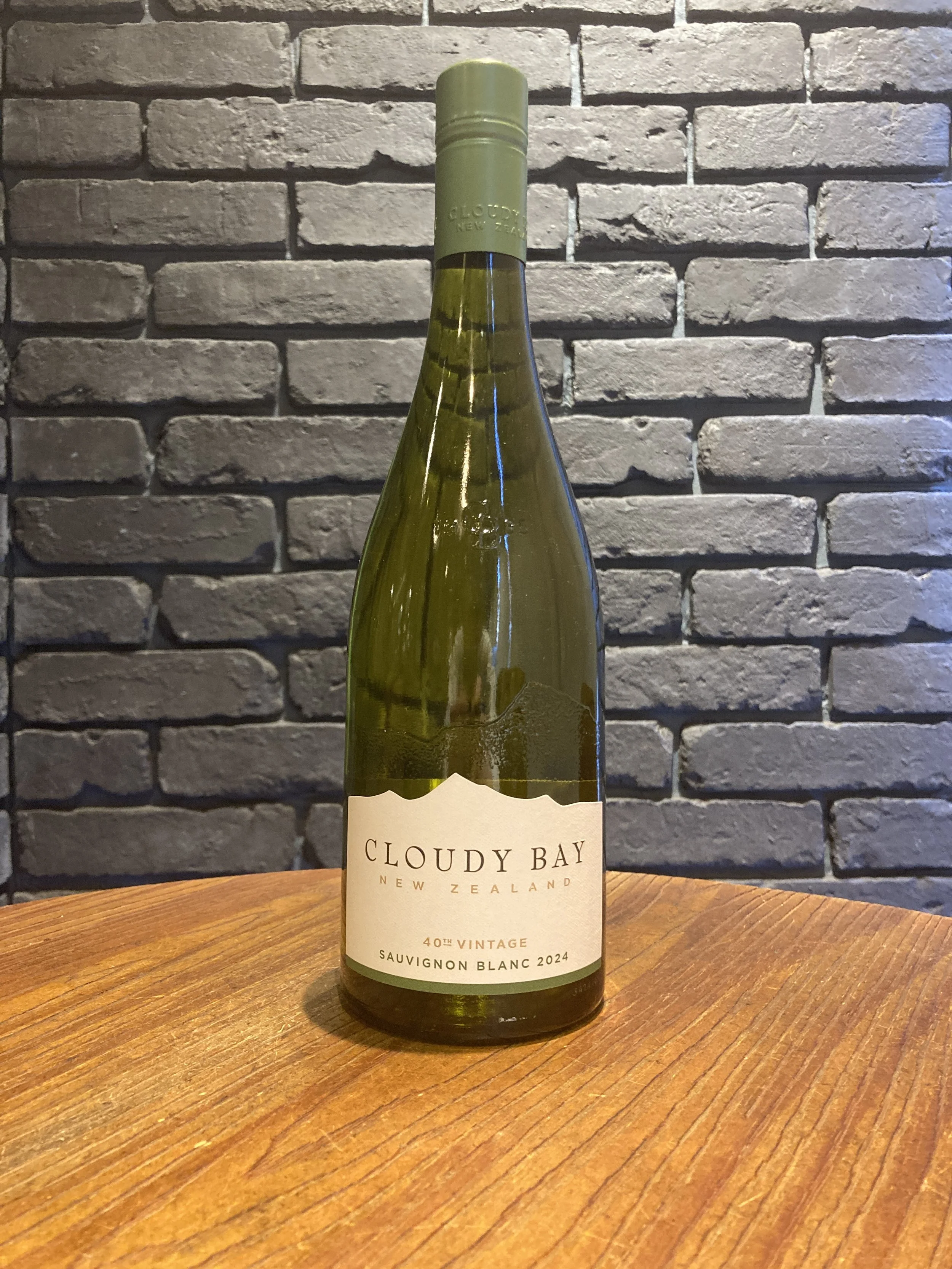 灰色のレンガ壁の前に置かれた緑色のワイン瓶。ラベルには「CLOUDY BAY NEW ZEALAND 40th VINTAGE SAUVIGNON BLANC 2024」と書かれている。木製のテーブルの上に置かれている。