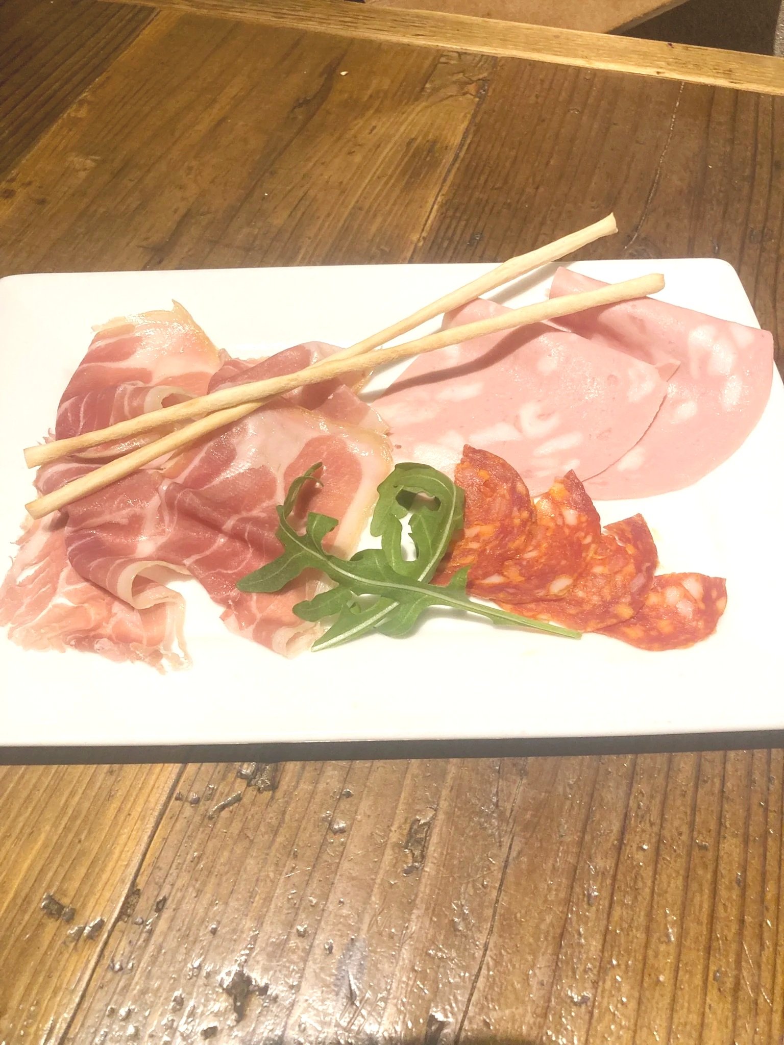 白い長方形の皿にサラミ、ハム、ローストビーフ、スパイシーソーセージとルッコラが盛り付けられている。長いクラッカー2本が上に置かれている。皿は木製テーブルの上に置かれている。