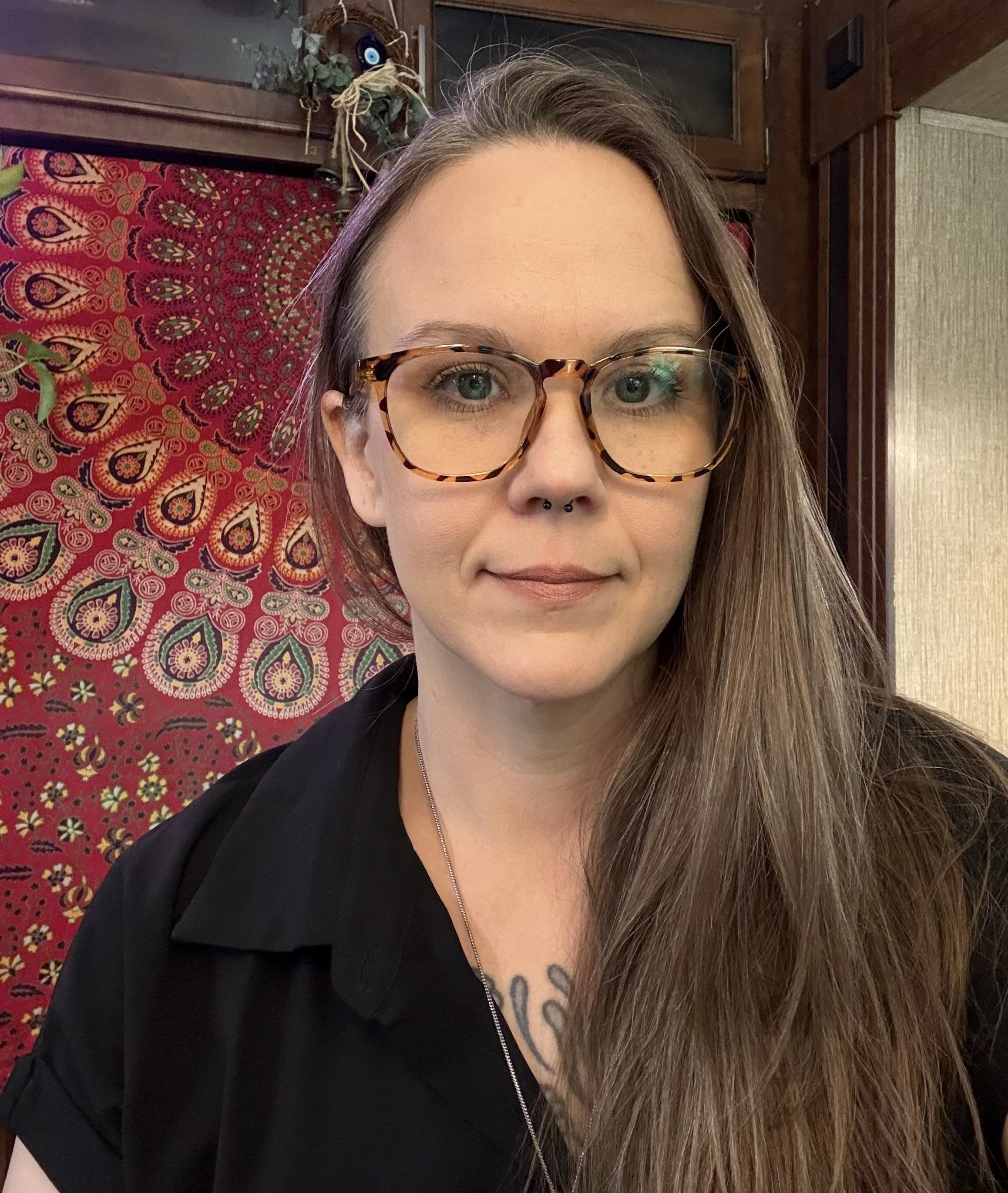 A woman with long brown hair, wearing tortoiseshell glasses and a septum piercing, smiling slightly in front of a colorful mandala-patterned fabric background.
