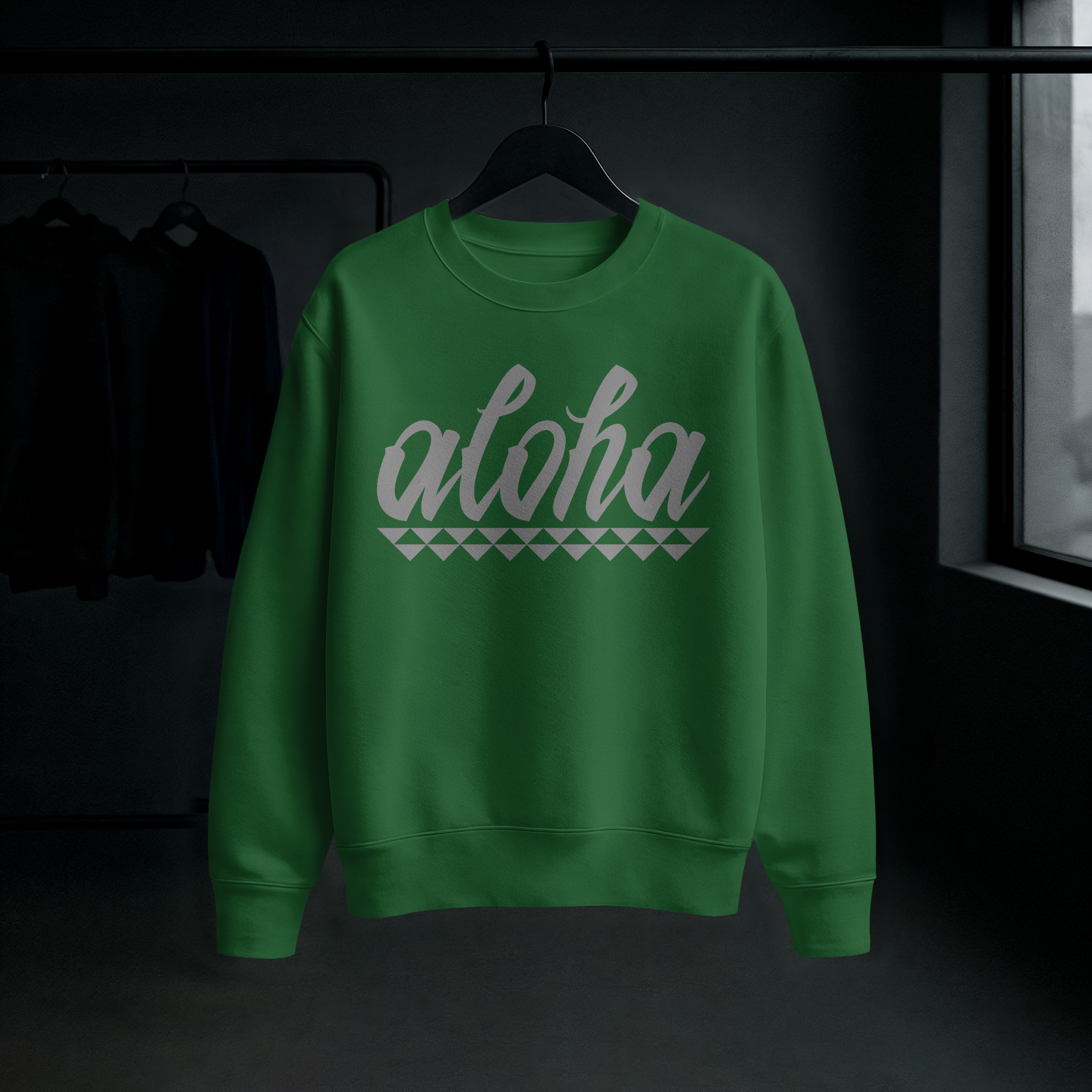 Green sweatshirt with the word 'Aloha' printed on the front, hanging on a hanger in a dark room.