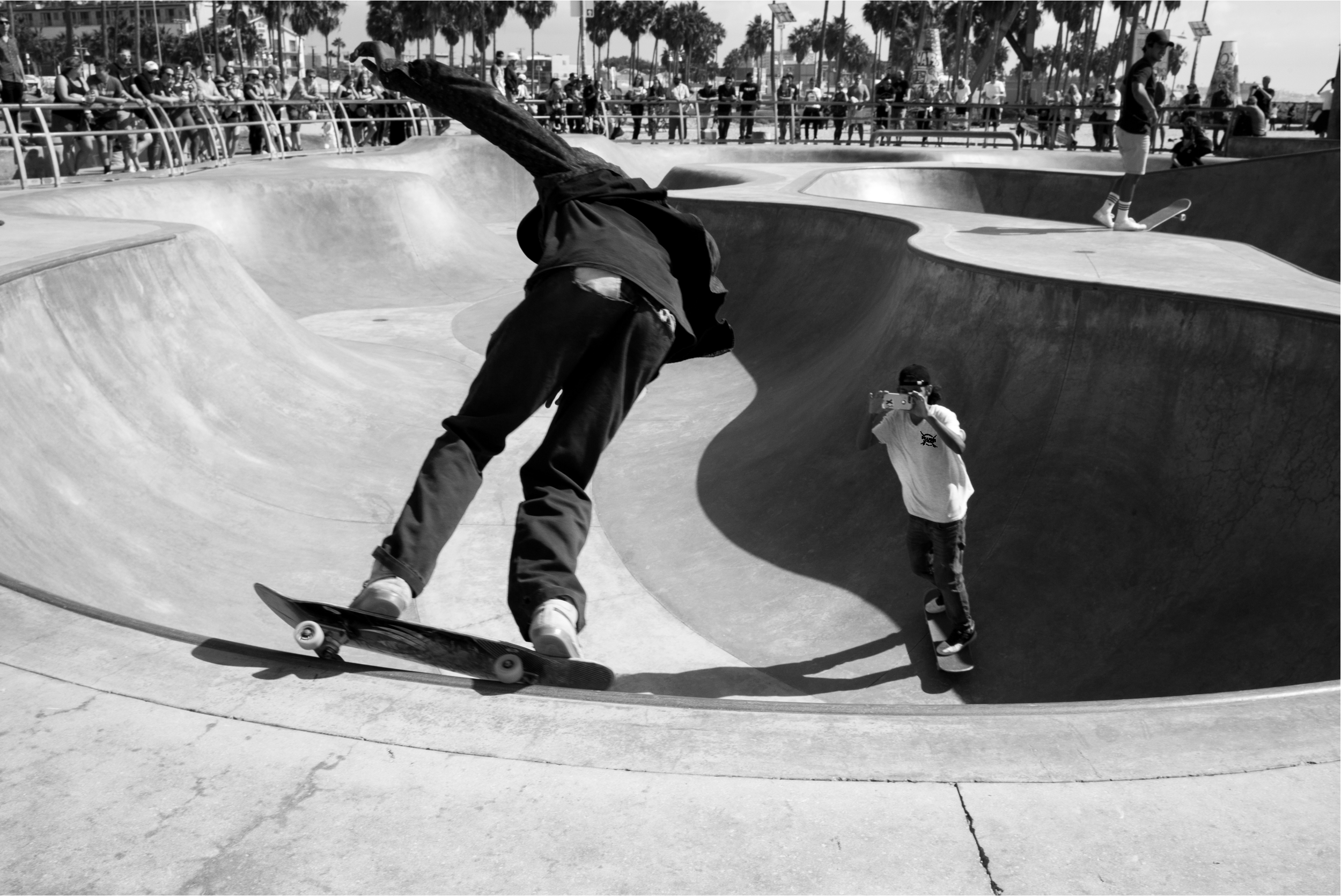 Skateboarder performing a trick in a skate park with spectators watching in the background, and a person taking a photo.