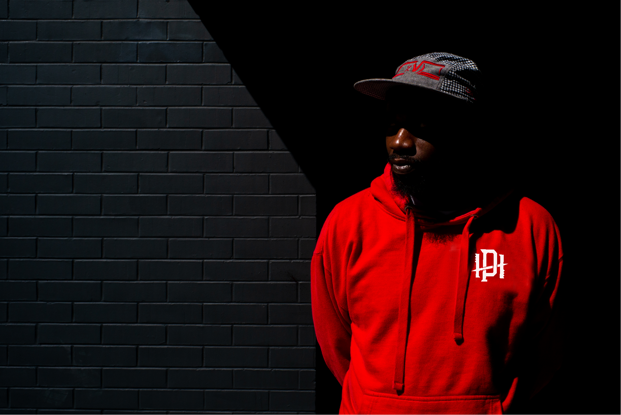 Man wearing a red hoodie and a black-and-white checkered cap with red accents stands against a black brick wall with shadows, partially obscured by darkness.