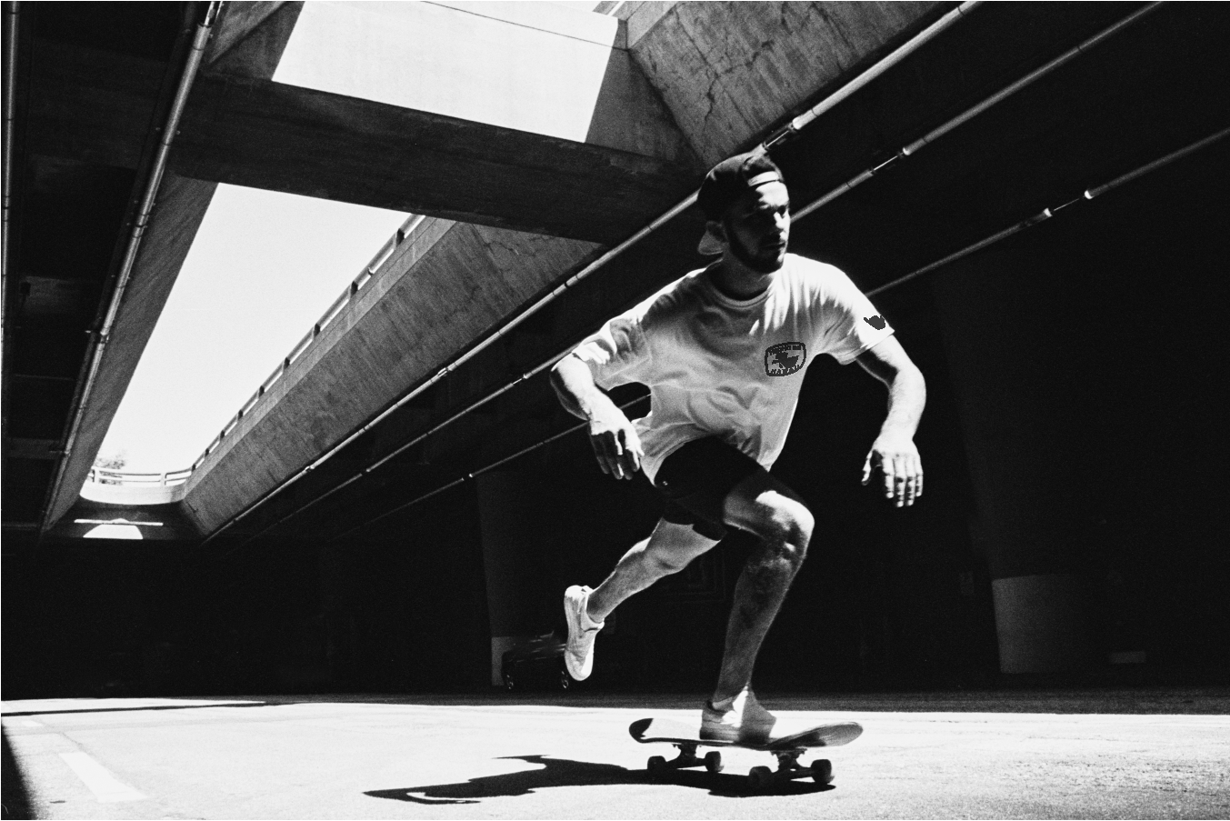 A young man skateboarding under a concrete bridge or overpass in an outdoor urban setting, shadowed area with sunlight illuminating the ground.