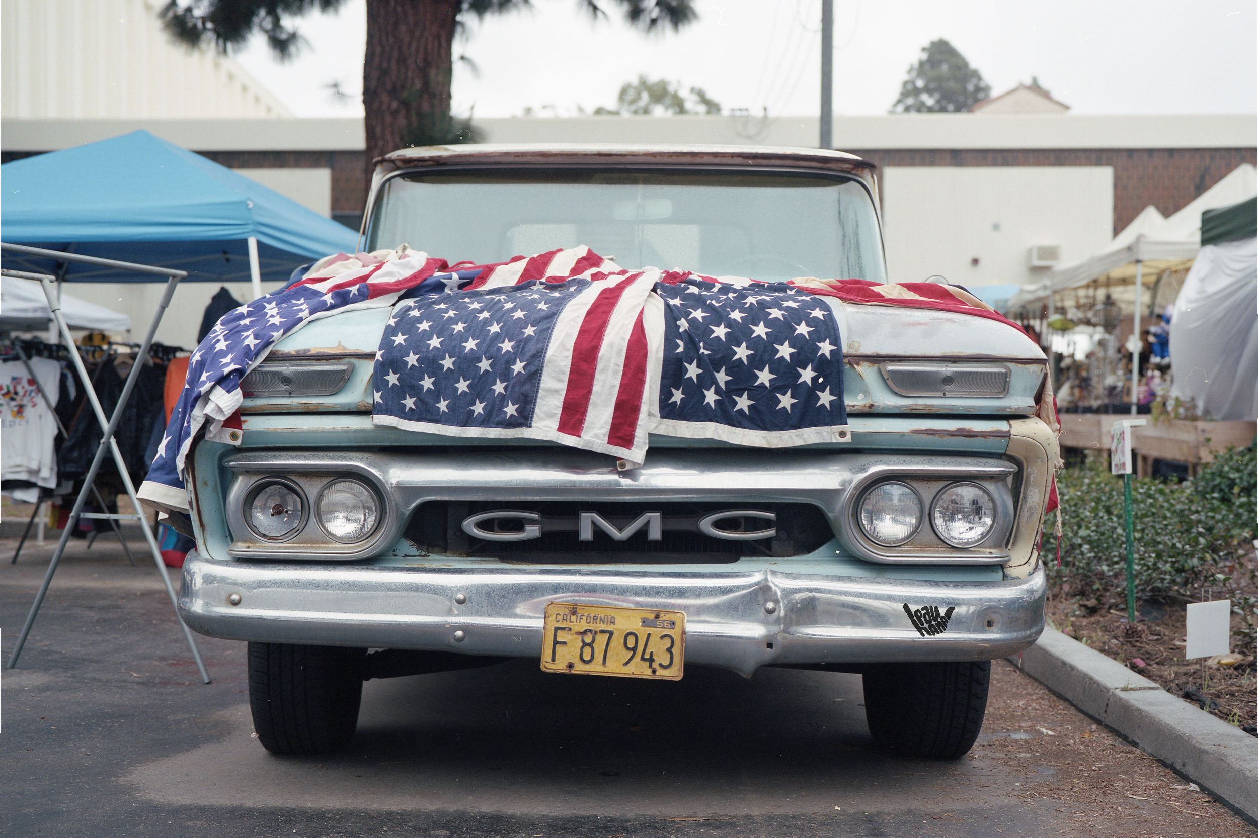 An old GMC truck with a rusted top, decorated with an American flag draped over the hood, parked in front of a market or fair with tents and other booths around.