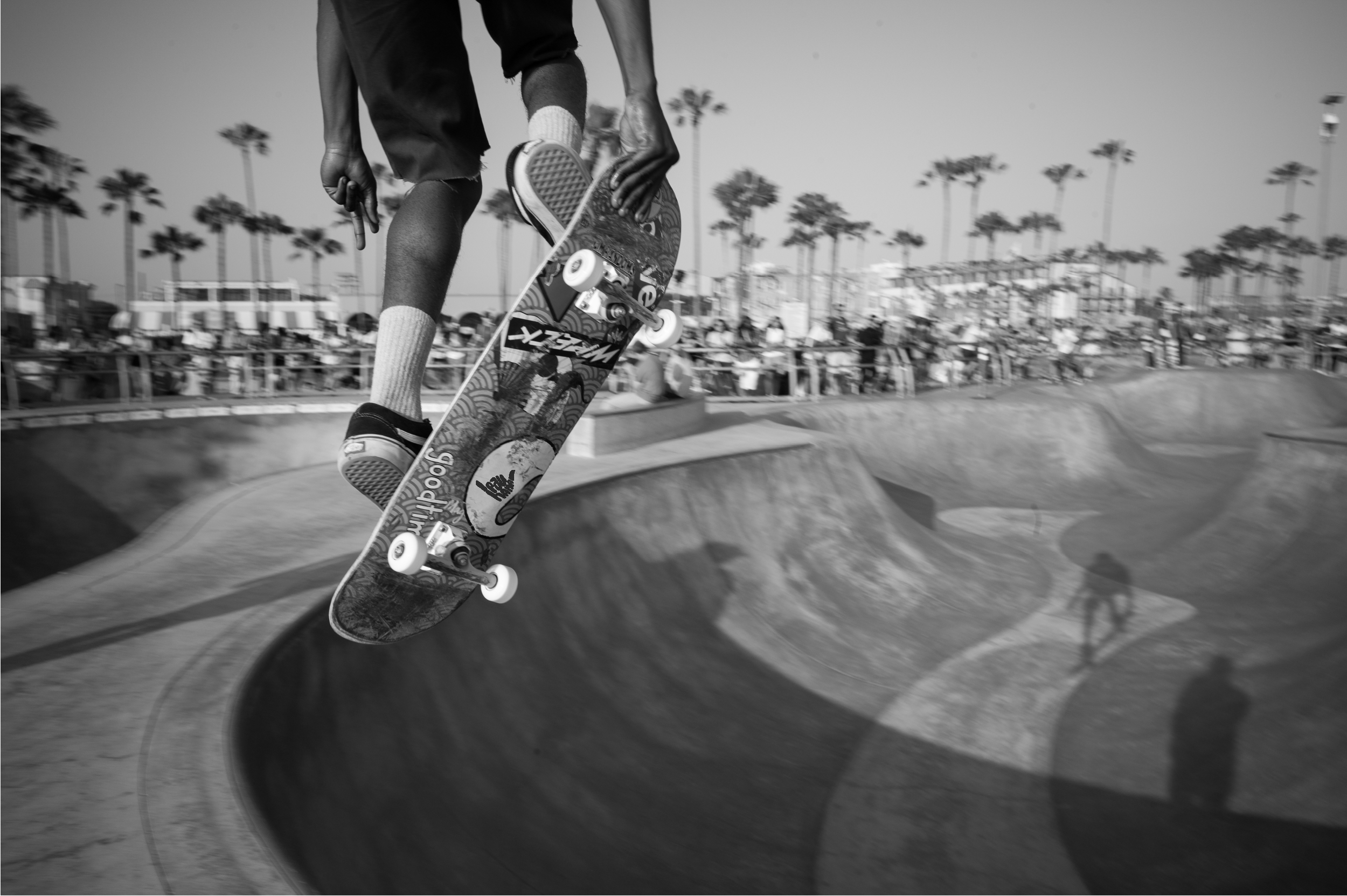 Skateboarder performing a jump over a skate bowl at a skate park with palm trees and spectators in the background.