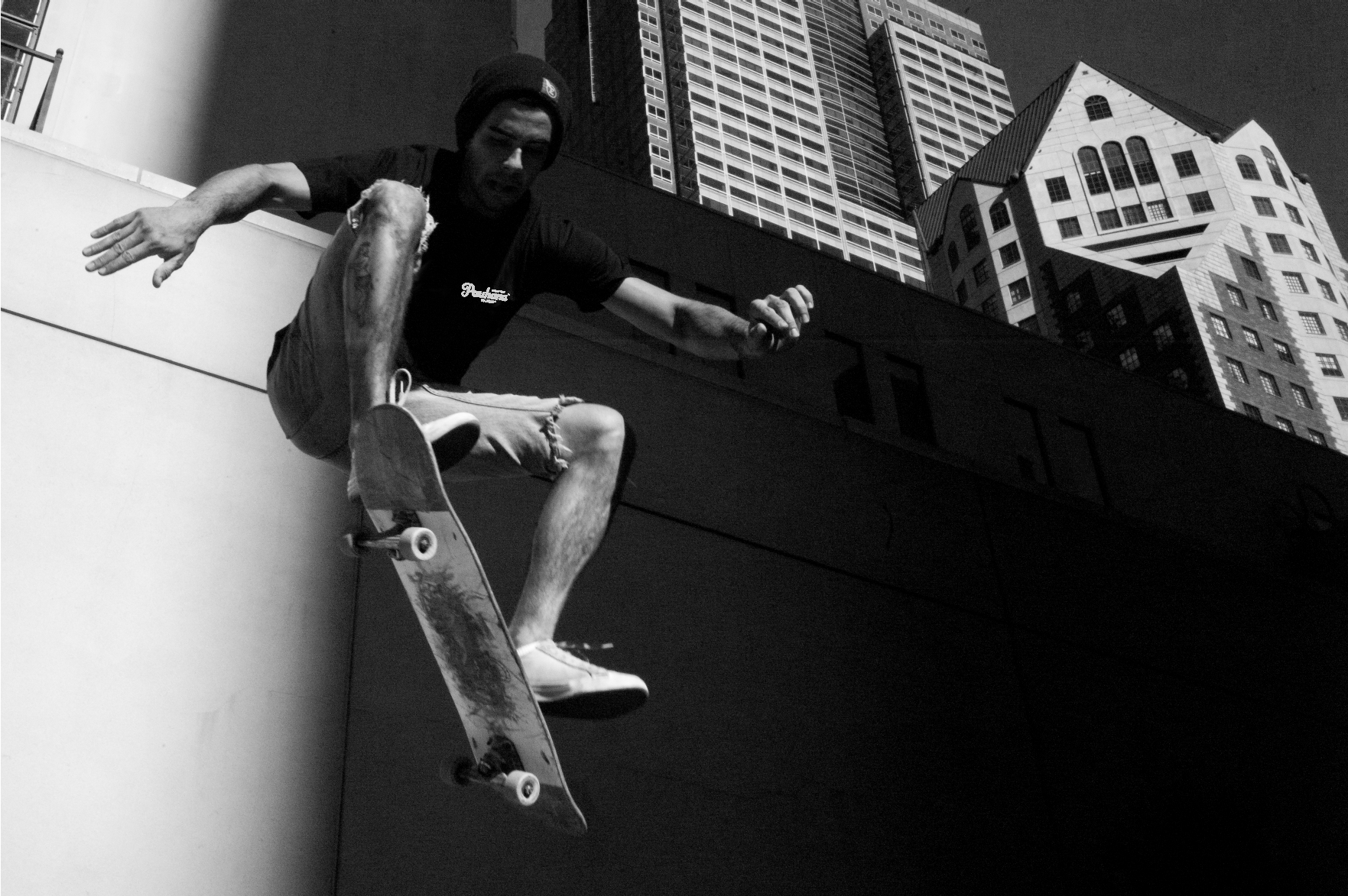 A black and white photo of a skateboarder mid-air performing an Ollie against an urban backdrop of tall buildings.