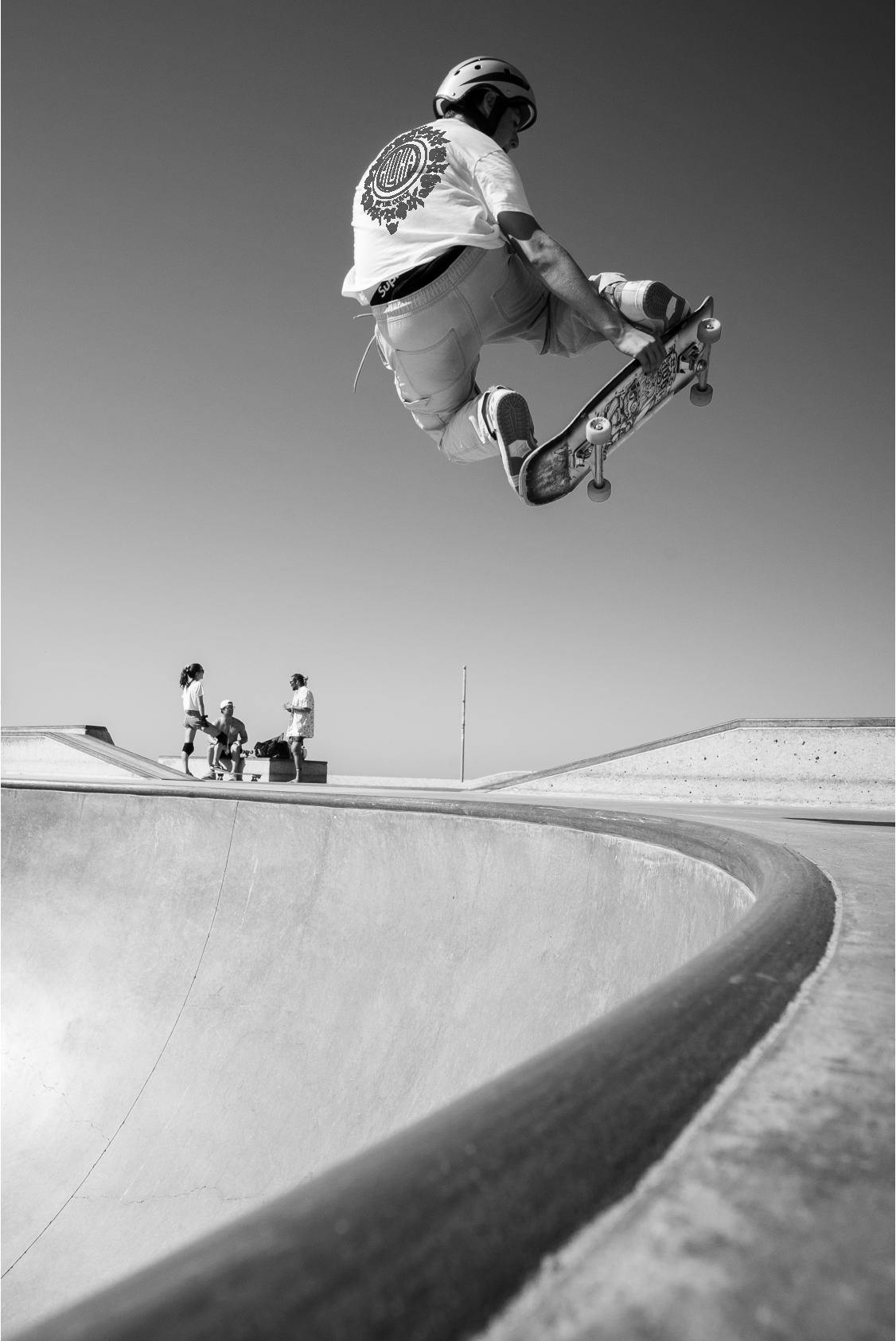 A skateboarder mid-air performing a trick at a skate park, with a few people in the background watching.