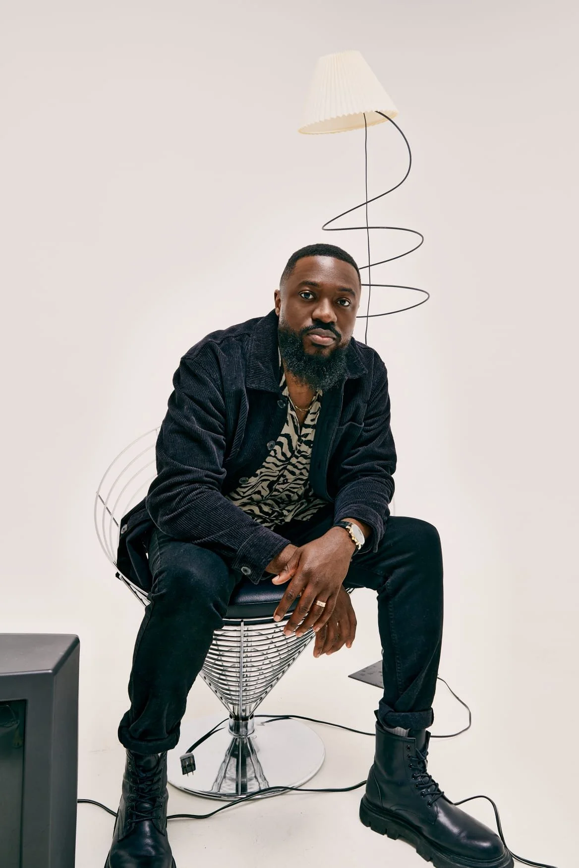 A man with a beard and short hair, wearing a black jacket, animal print shirt, black pants, and black boots, sitting on a modern wire chair in front of a plain white background. A tall floor lamp with a pleated shade is behind him.