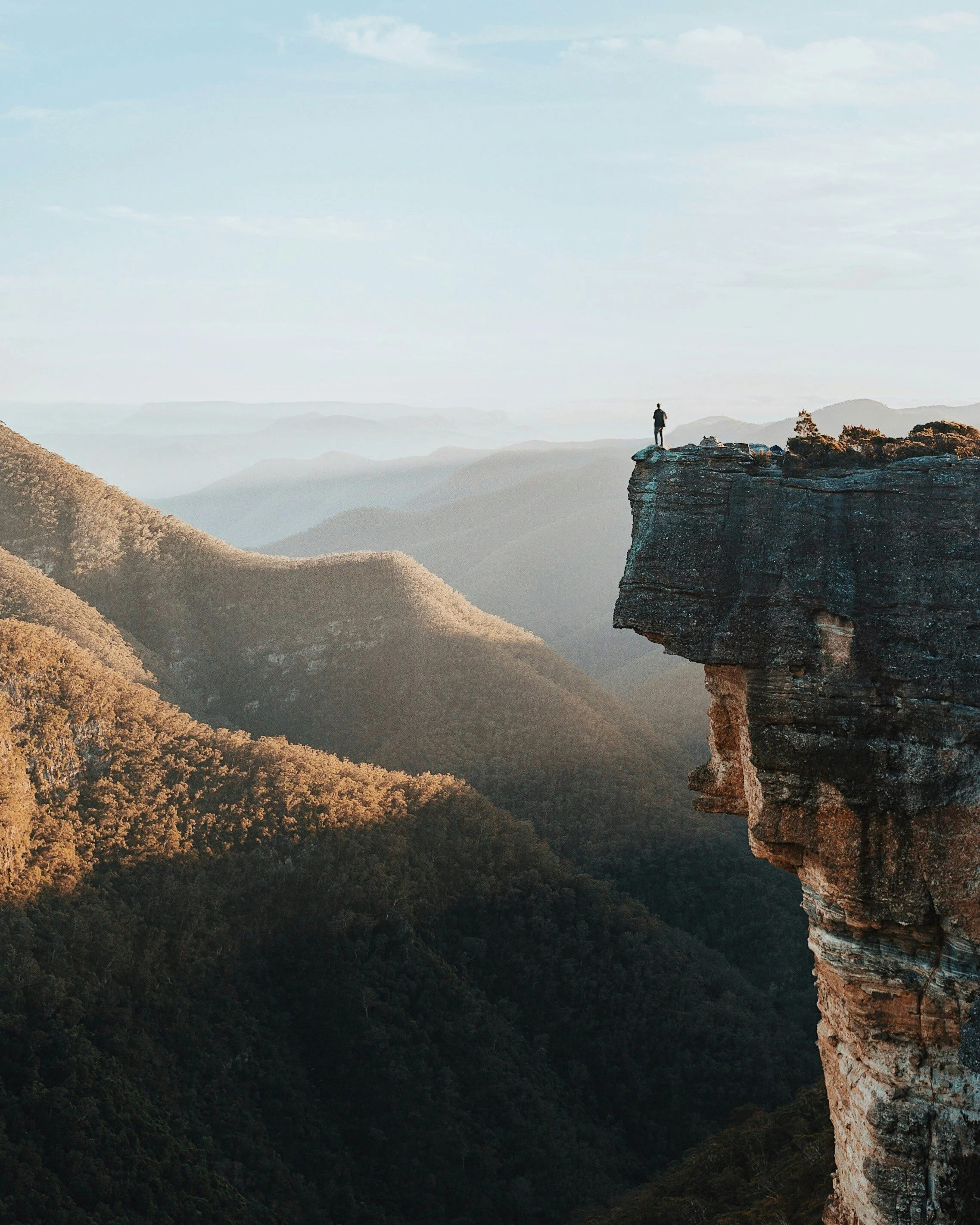 A person standing on the edge of a rocky cliff overlooking a mountain landscape at sunrise or sunset.