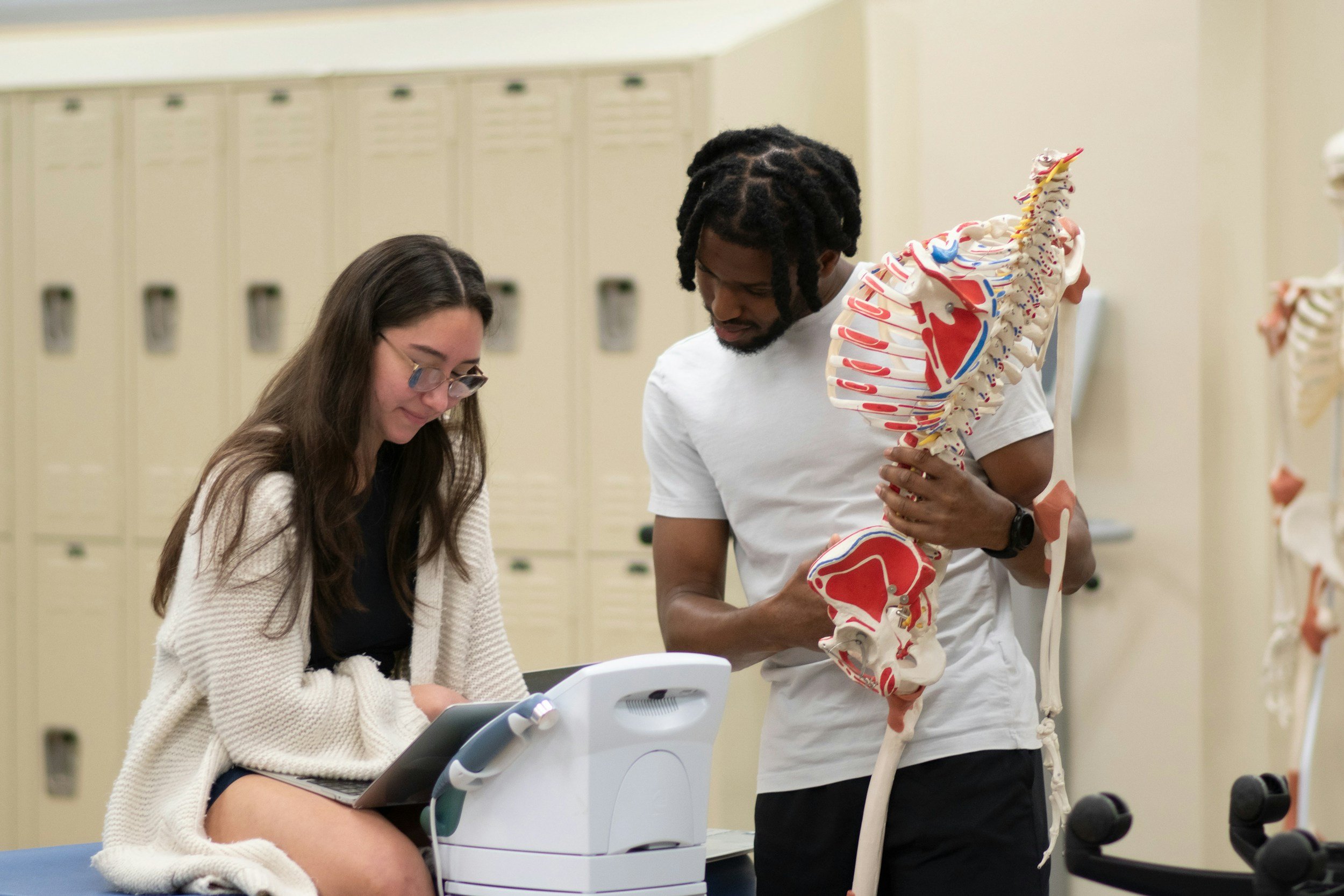 A young woman with glasses and long brown hair sitting in a classroom or lab, looking at a computer or medical device, while a young man with dreadlocks, wearing a white t-shirt, holding a detailed anatomical model of the human spine and rib cage, stands nearby in front of beige lockers.