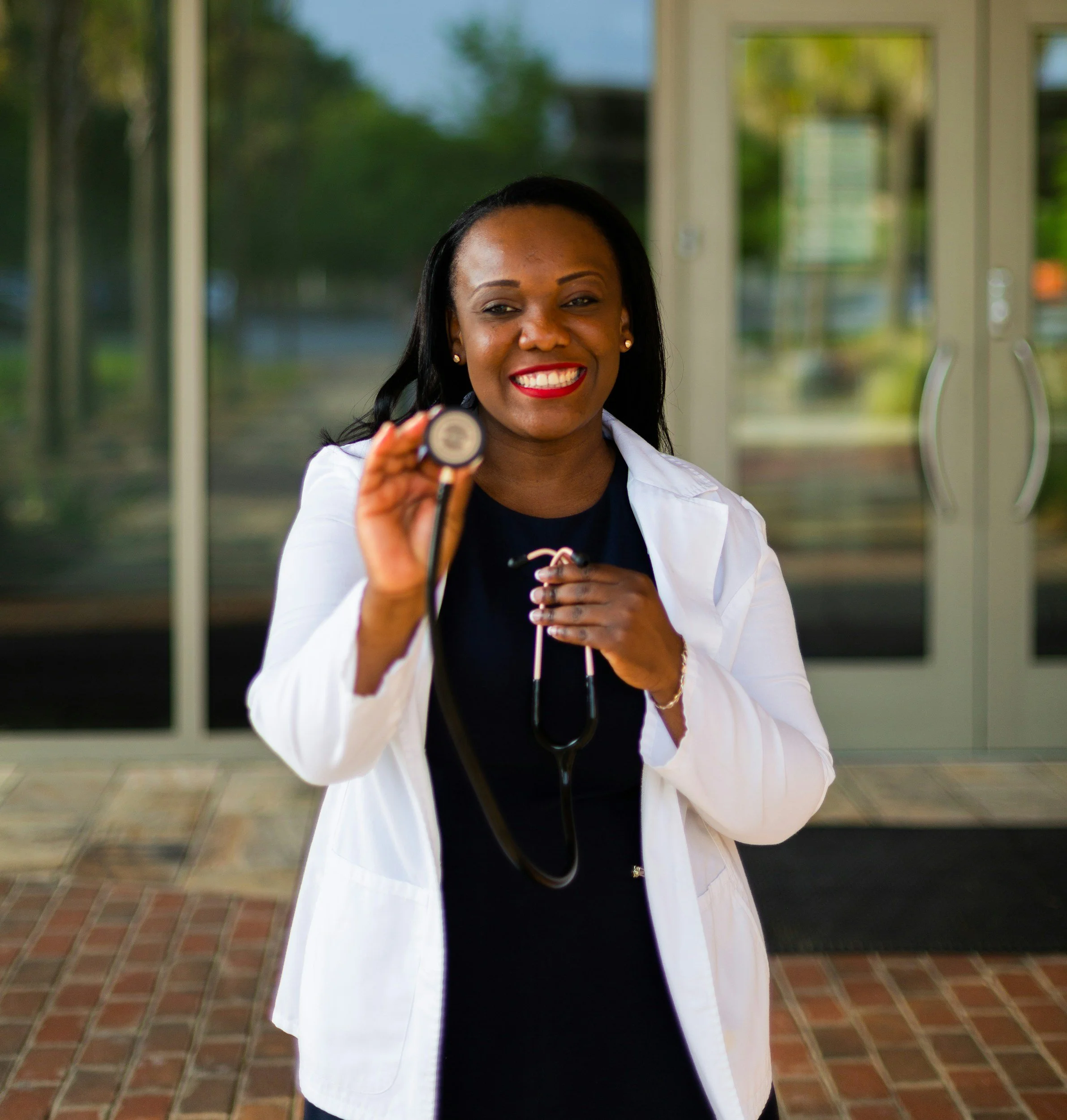 A smiling female healthcare worker holding a stethoscope outside a building.
