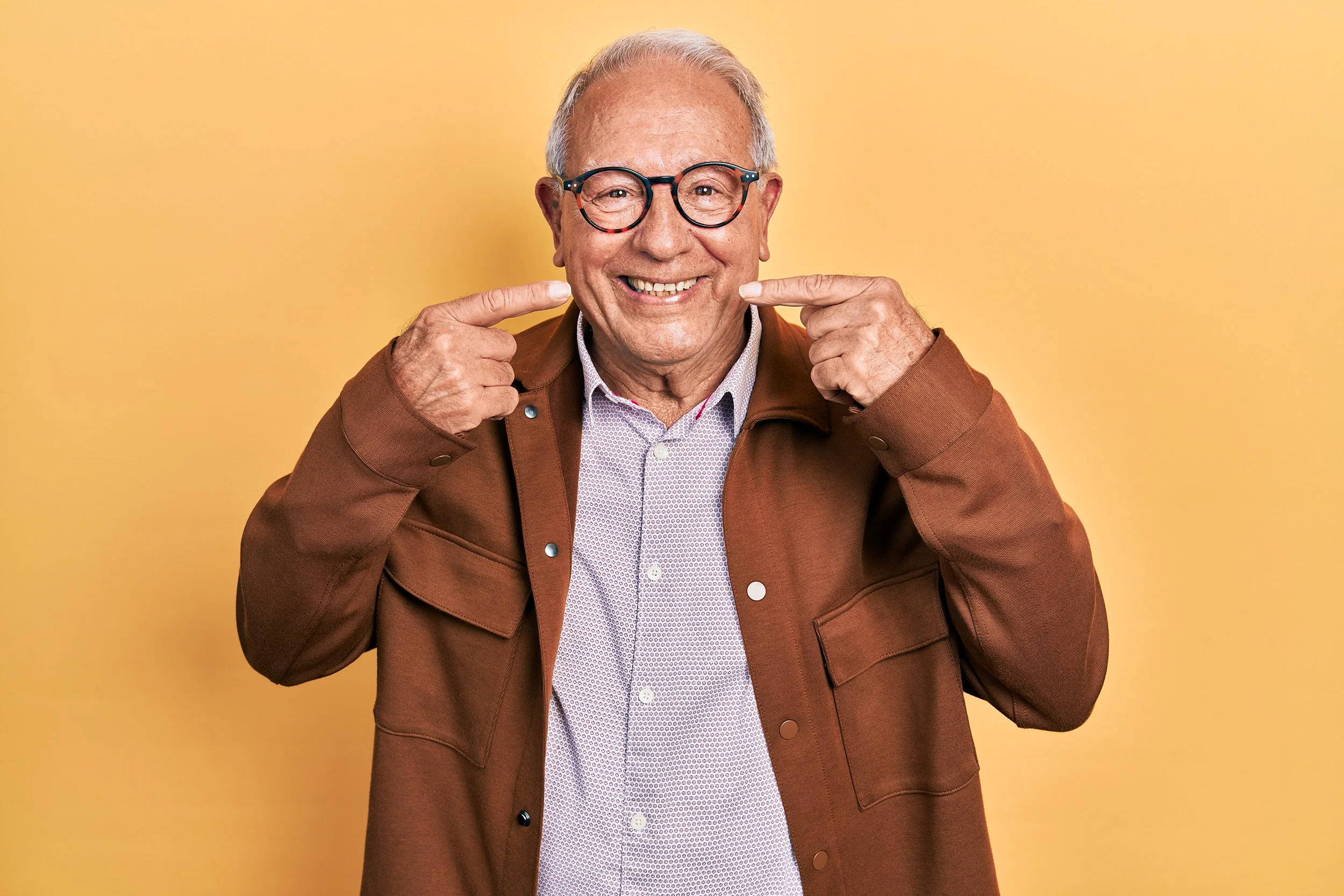 Smiling older man with glasses pointing at his smile, wearing a brown jacket and light shirt, standing against a yellow background.