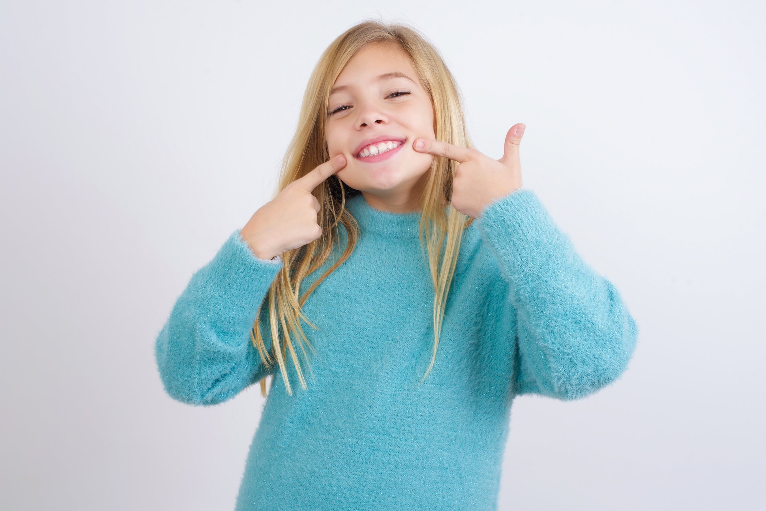Young girl in a blue sweater smiling and pointing to her teeth with both index fingers.