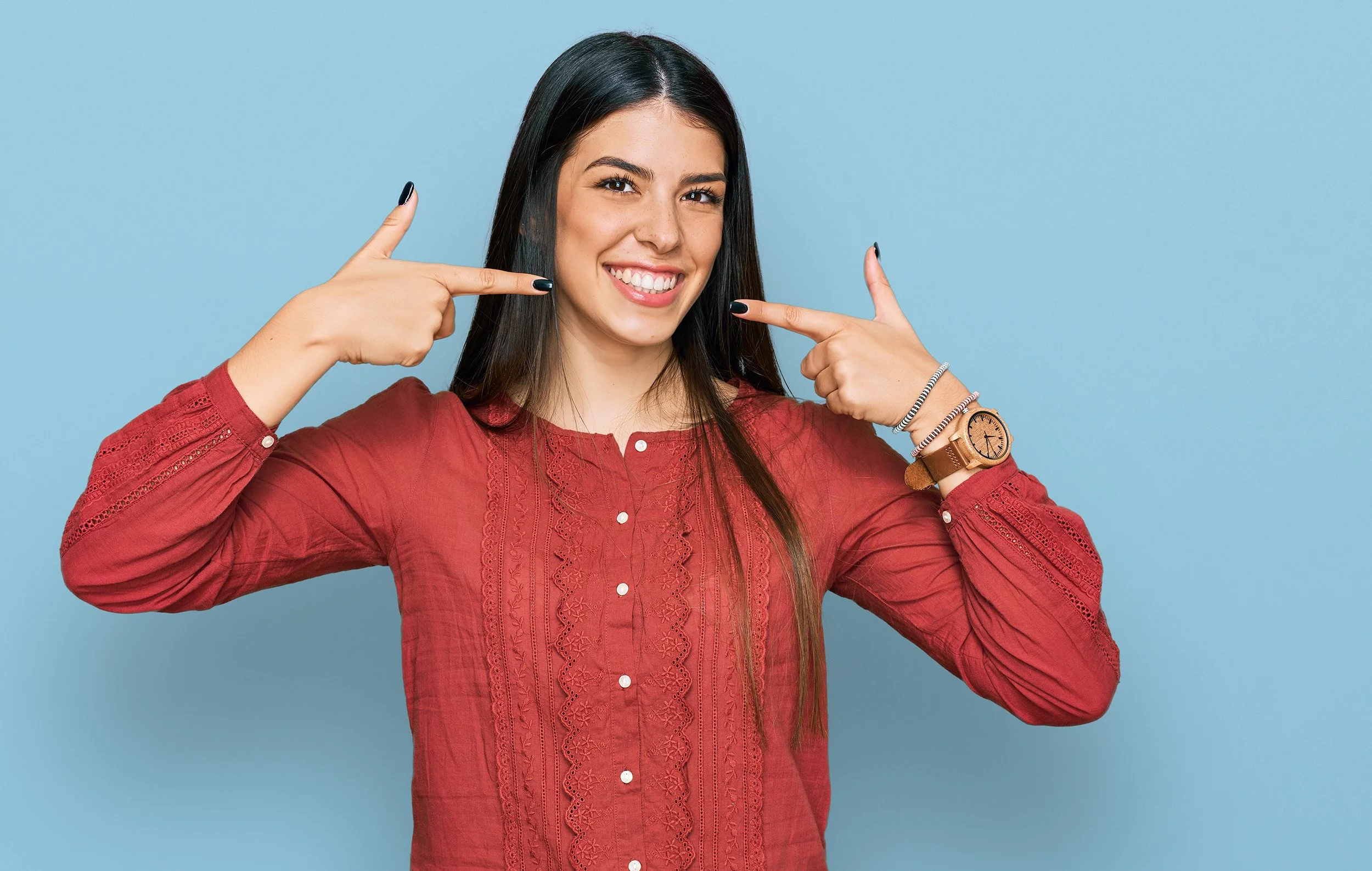 Young woman in a red blouse smiling and pointing at her teeth.