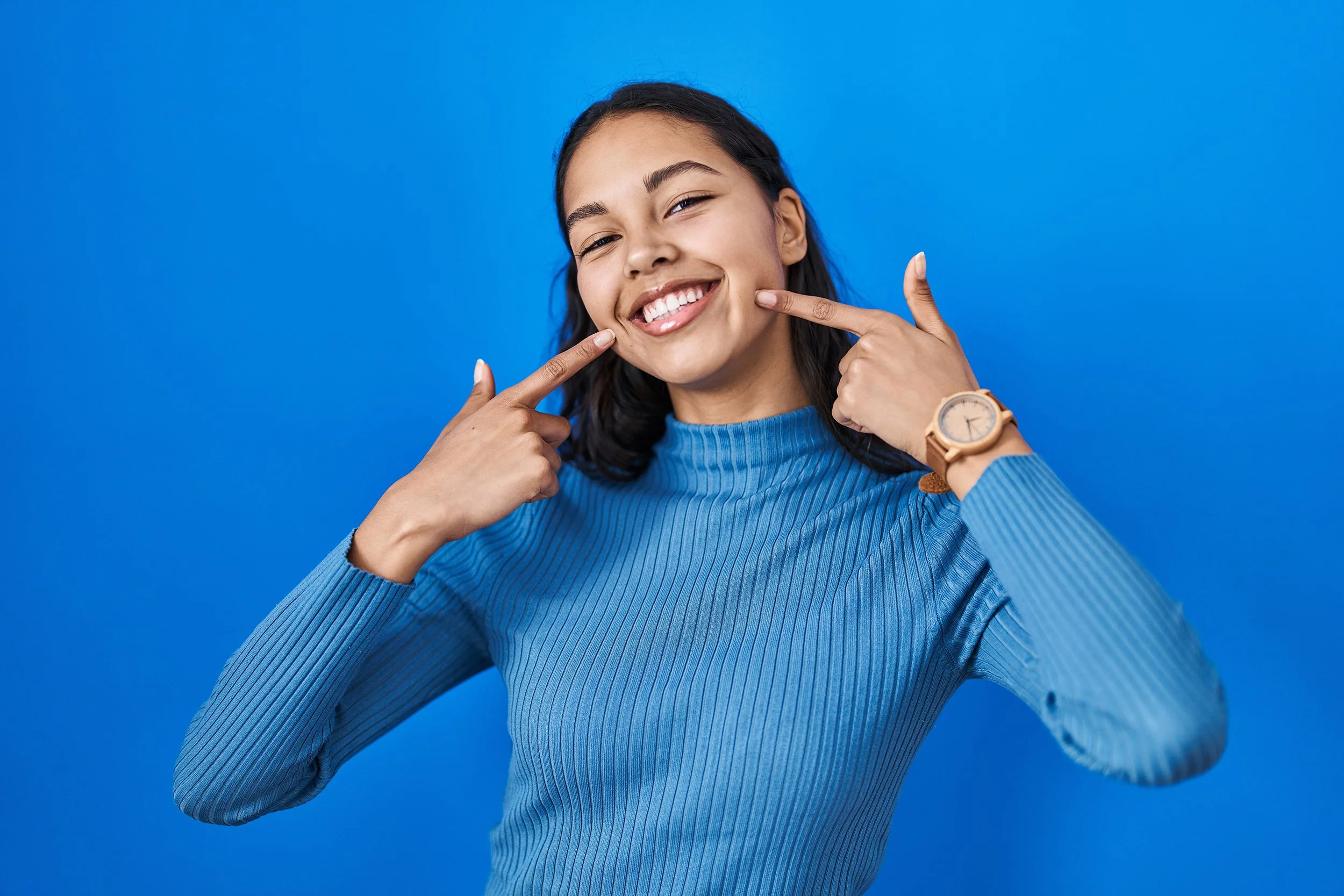 Woman in blue sweater smiling and pointing to her cheeks against a blue background.