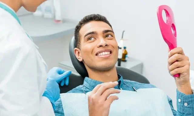 A dentist wearing gloves talks to a smiling patient holding a red hand mirror.
