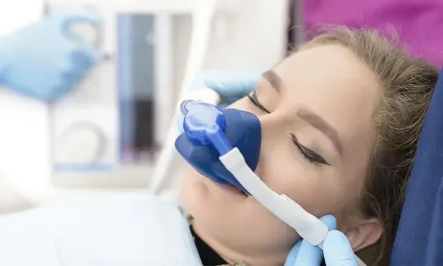 Patient wearing a blue nasal mask with eyes closed, possibly receiving sedation or anesthesia, attended by a healthcare professional wearing gloves.