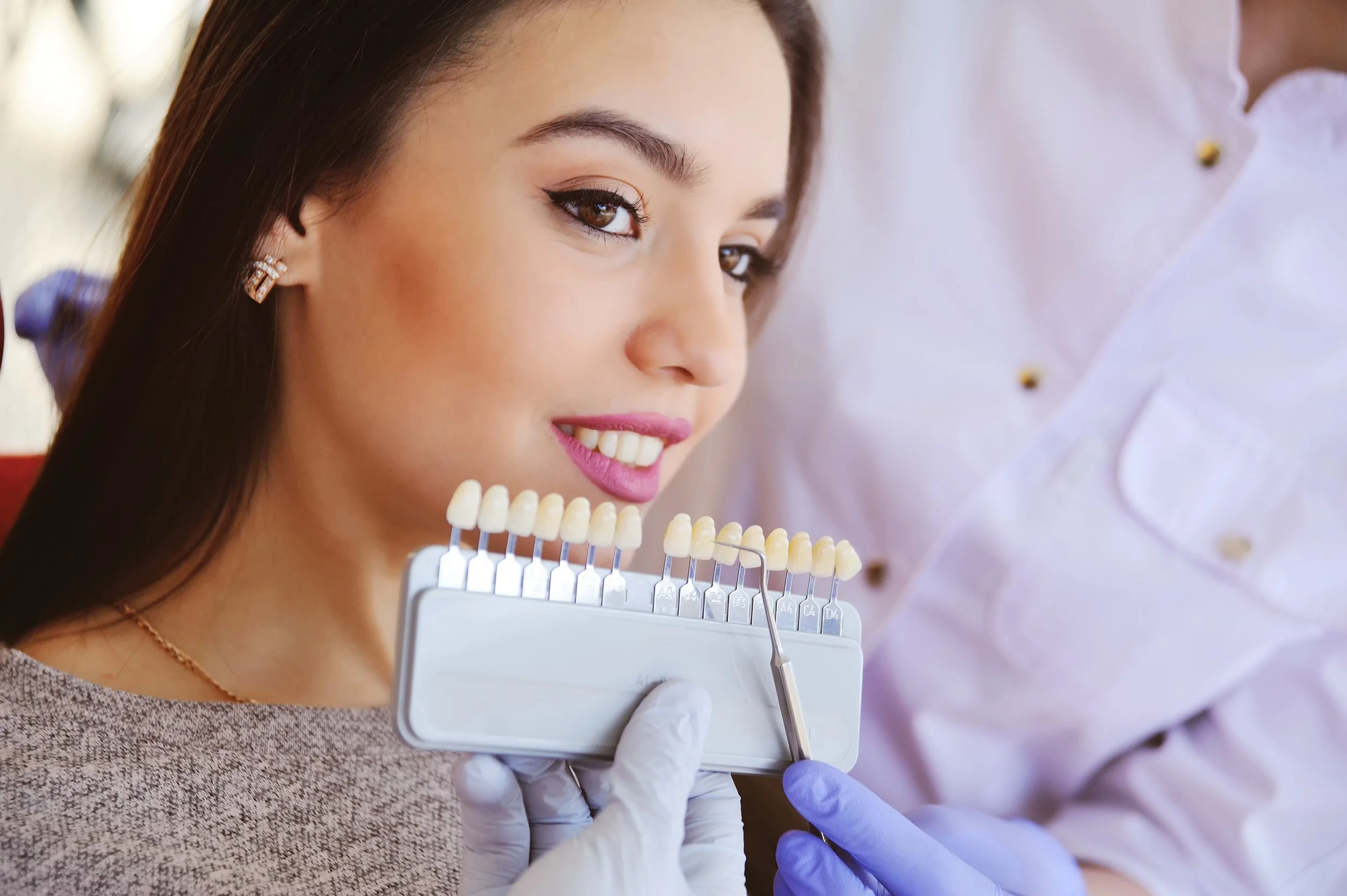 A woman with long dark hair and makeup is smiling at a dentist or dental technician, who is holding a shade guide with multiple teeth samples for color matching.