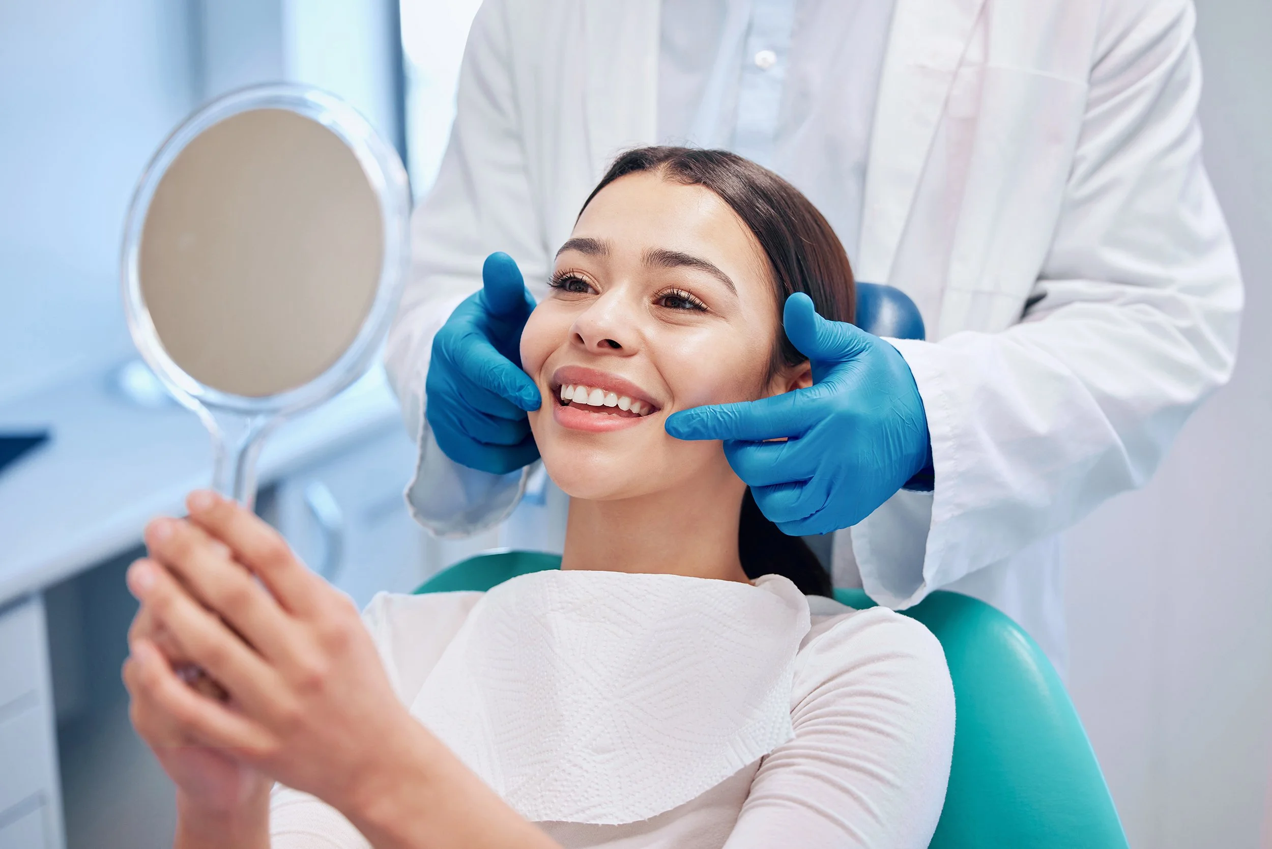 Woman seated in dental chair smiling while a dentist examines her teeth with gloved hands in a dental clinic.