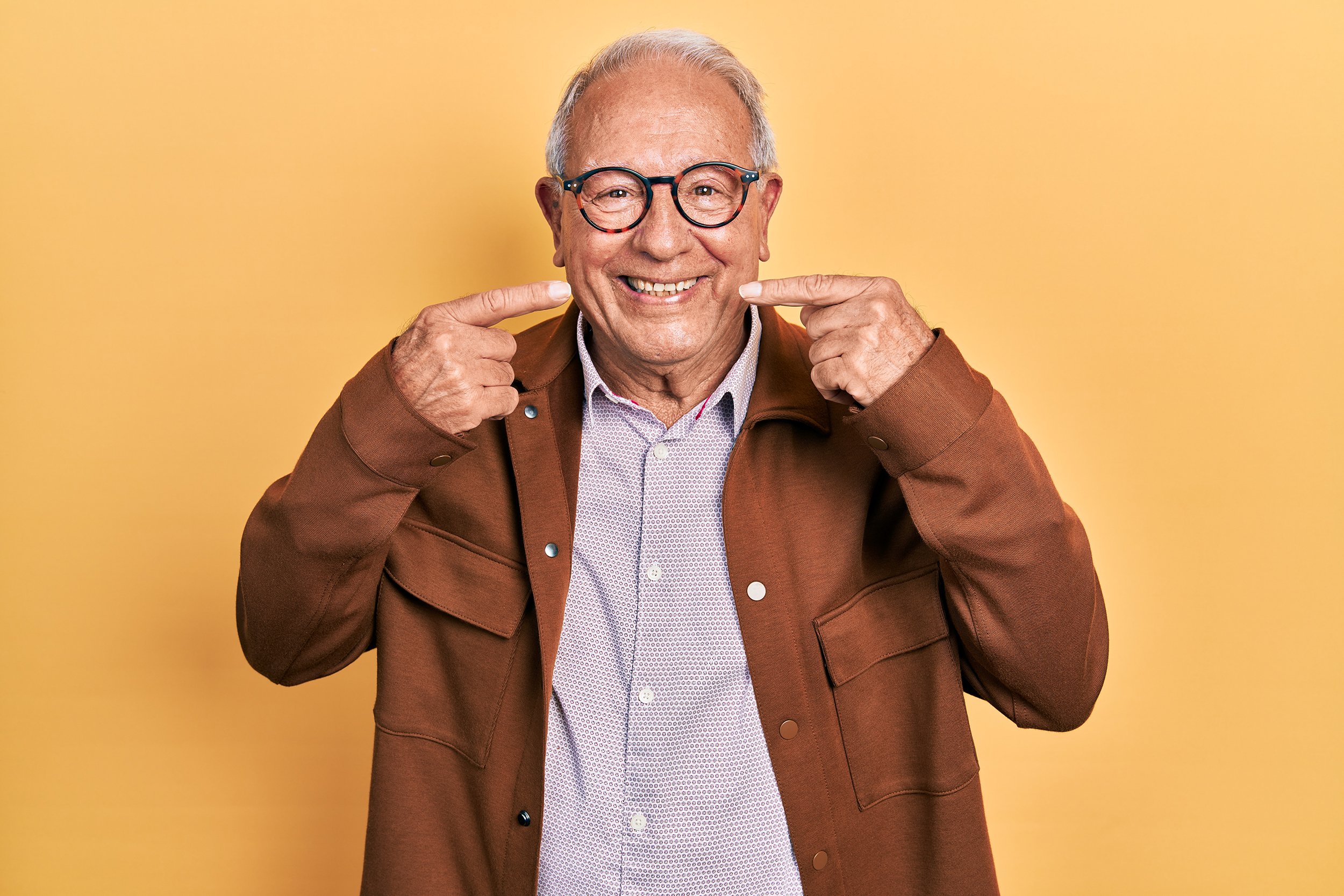 Elderly man smiling and pointing at his smile, wearing glasses and a brown jacket against a yellow background.