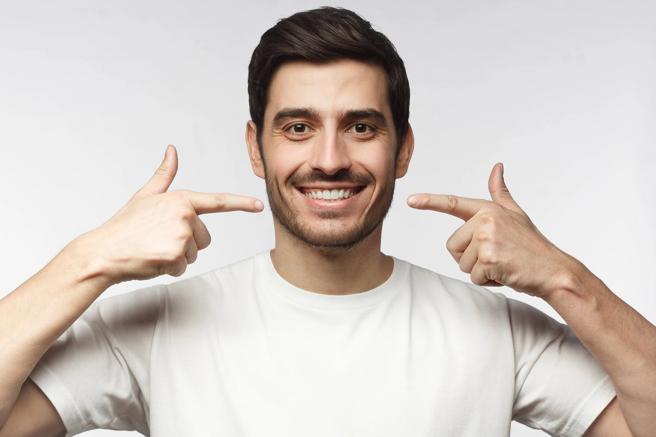 Happy young man pointing at his head with both index fingers, smiling, wearing a white t-shirt, against a plain light background.