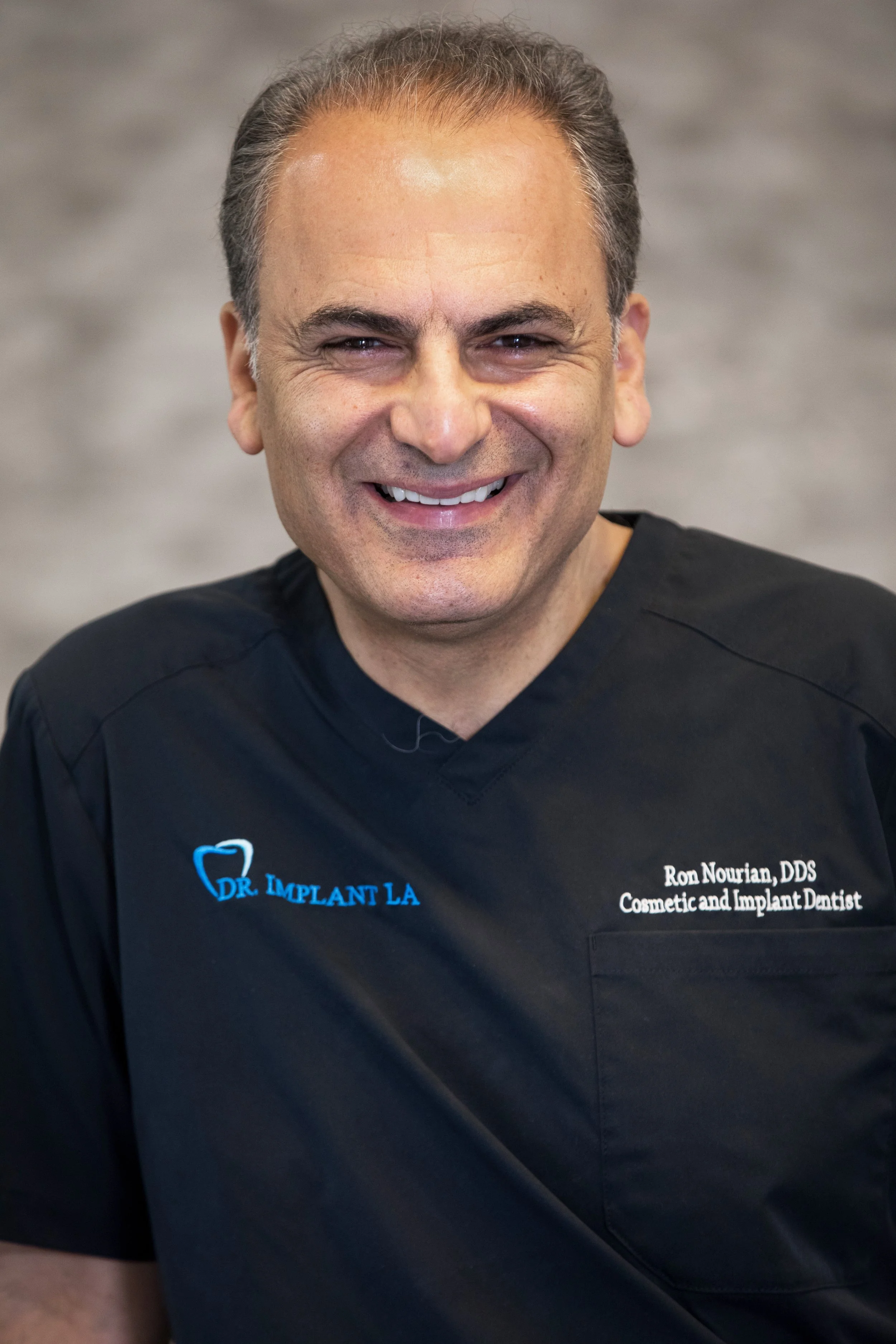 A smiling man in black medical scrubs with blue and white text, standing against a blurred neutral background.