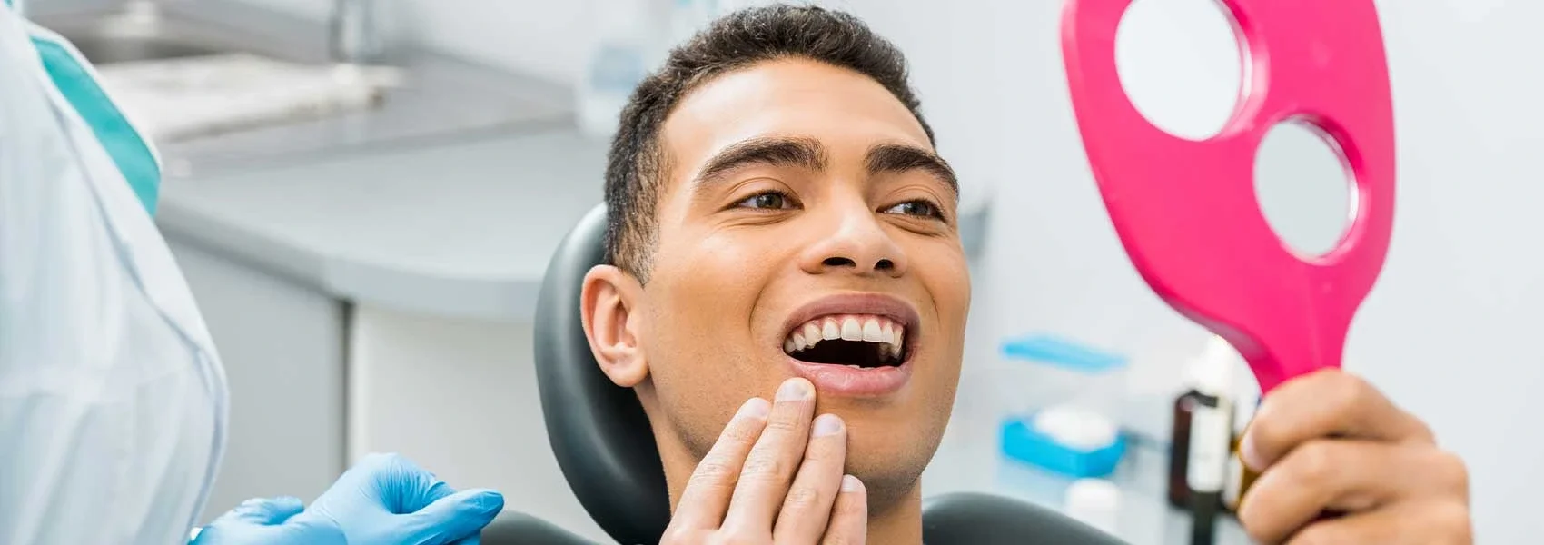 A young man at the dentist's office looking in a mirror, touching his chin, with the dentist's gloved hand near his face. The man is smiling and appears to be examining his teeth.