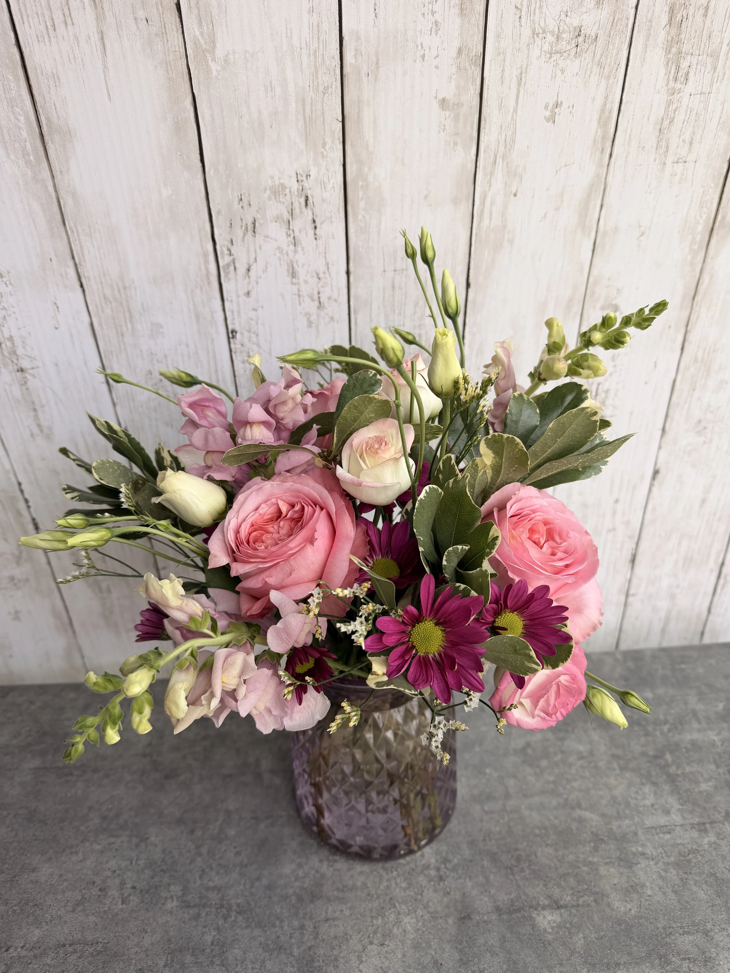 A floral arrangement with pink roses, purple daisies, white lisianthus, and greenery in a textured purple glass vase on a gray surface with a white wooden background.