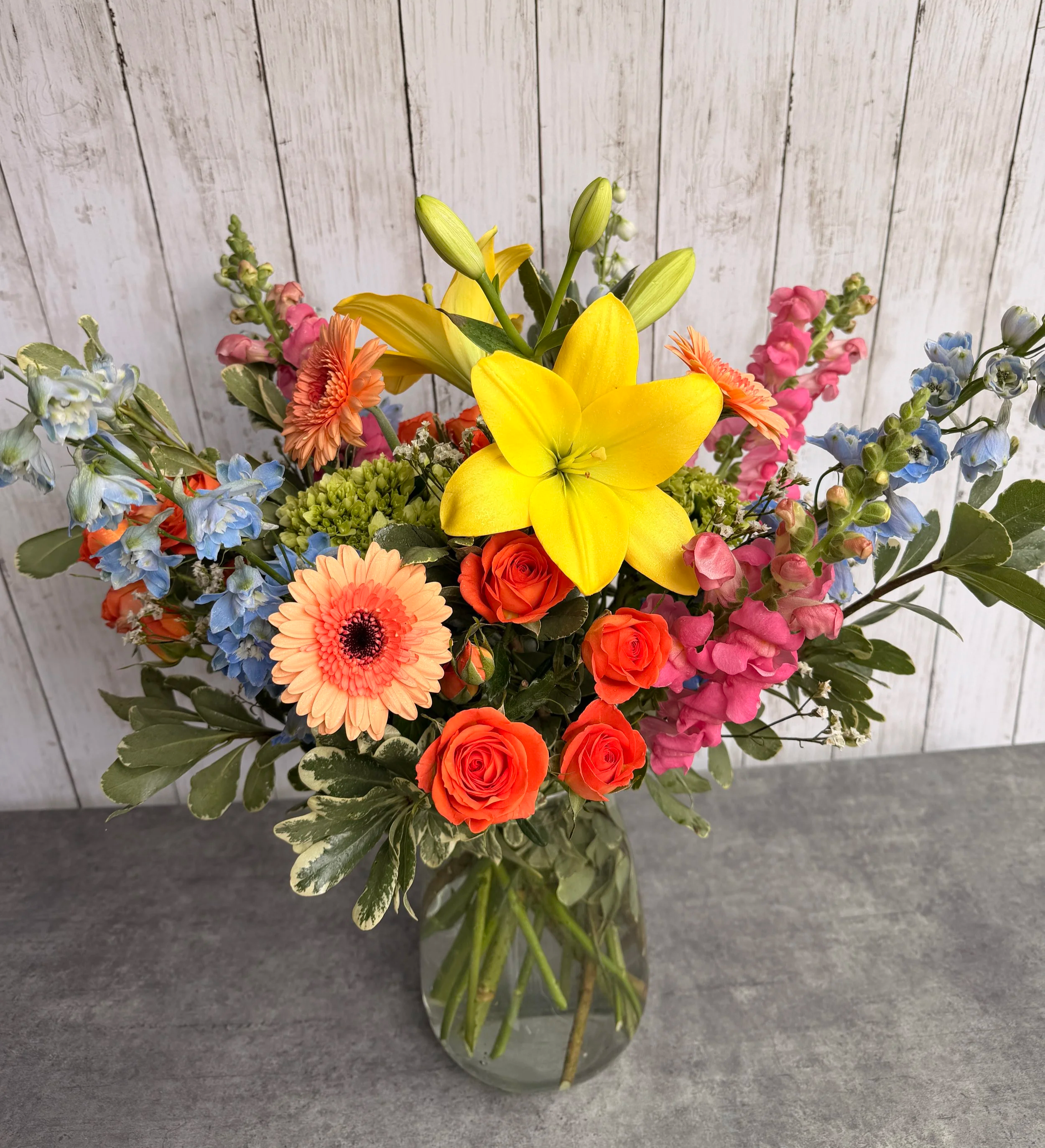 A colorful bouquet of flowers in a glass vase, featuring yellow lilies, peach gerbera daisies, orange roses, pink snapdragons, blue delphiniums, and other mixed greenery, set against a white wooden background.