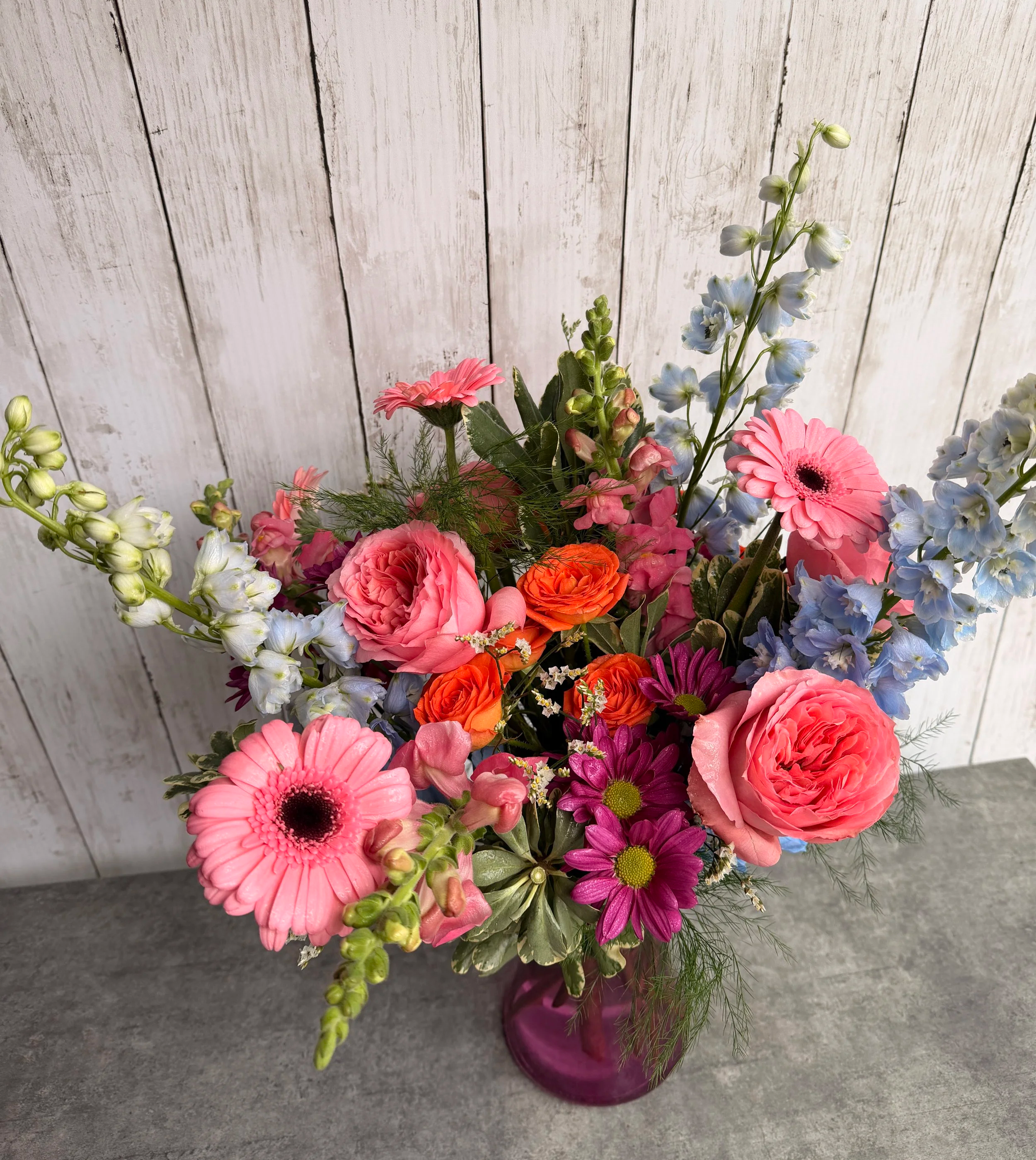 A colorful bouquet of flowers in a pink vase, featuring pink gerbera daisies, pink roses, orange ranunculus, purple daisies, blue delphiniums, and greenery, set against a white wooden background.