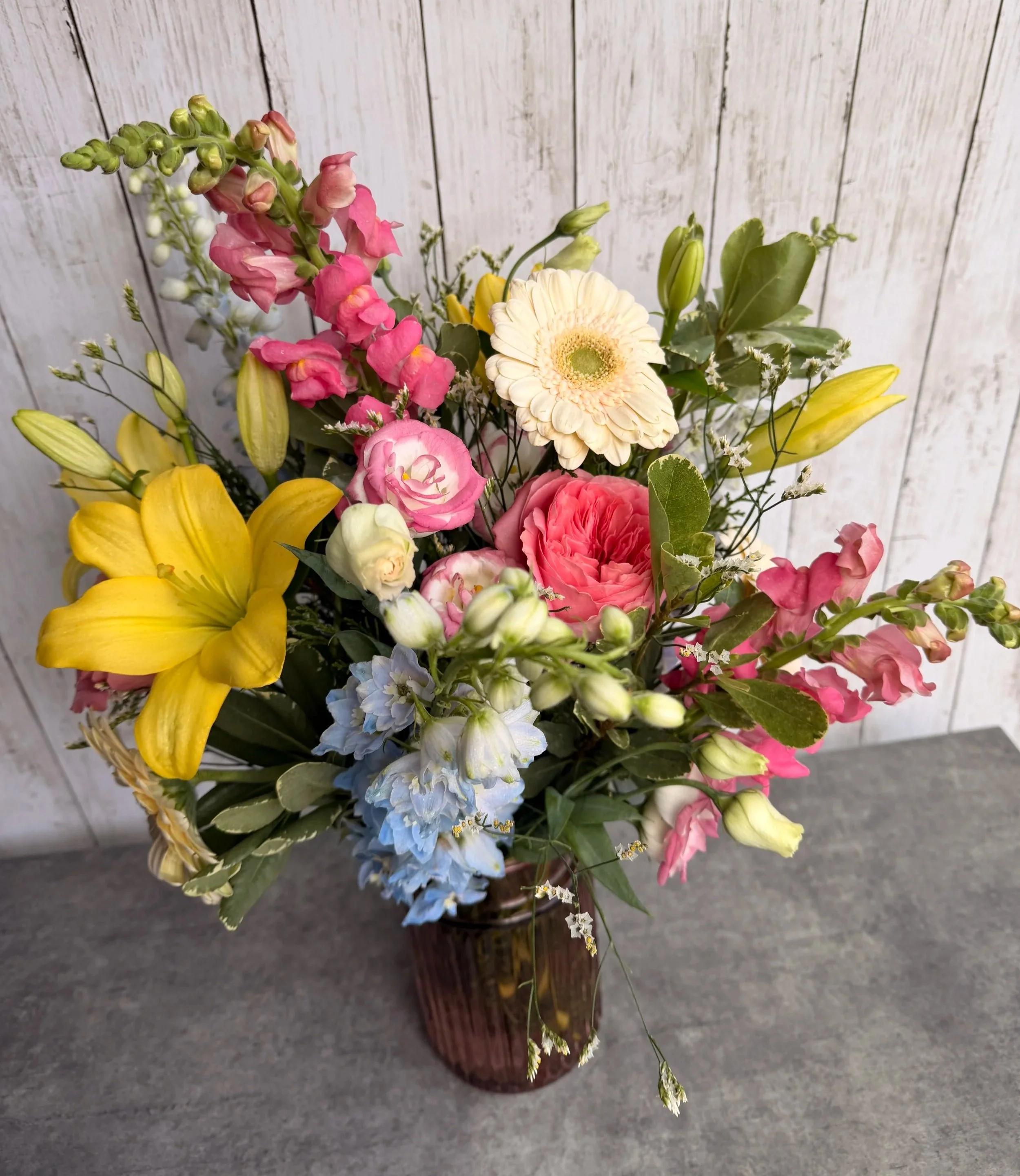 A colorful bouquet of various flowers in a brown vase on a gray surface against a white wooden panel background.
