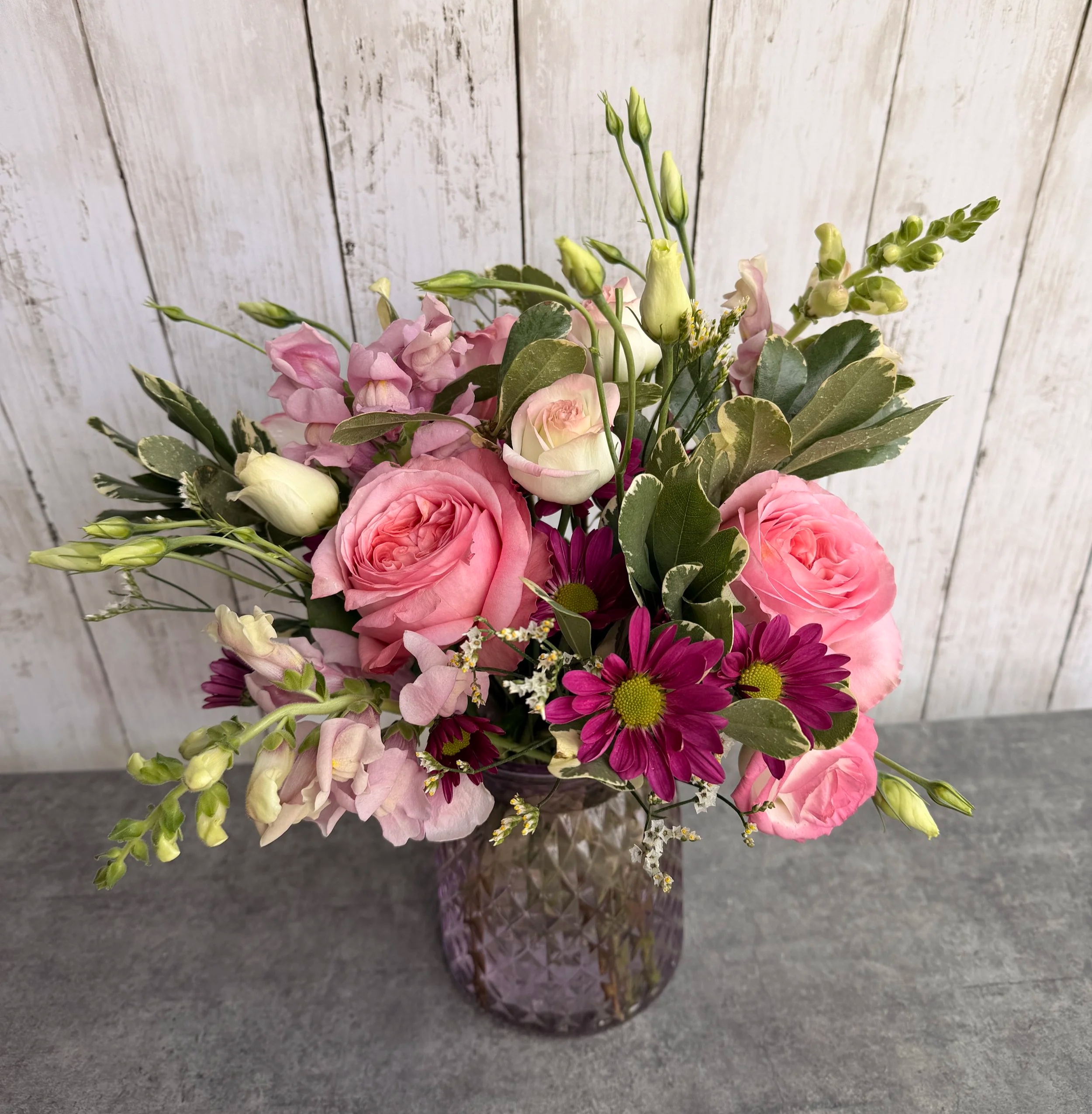 A floral arrangement with pink roses, purple daisies, white lisianthus, and greenery in a textured purple glass vase on a gray surface with a white wooden background.
