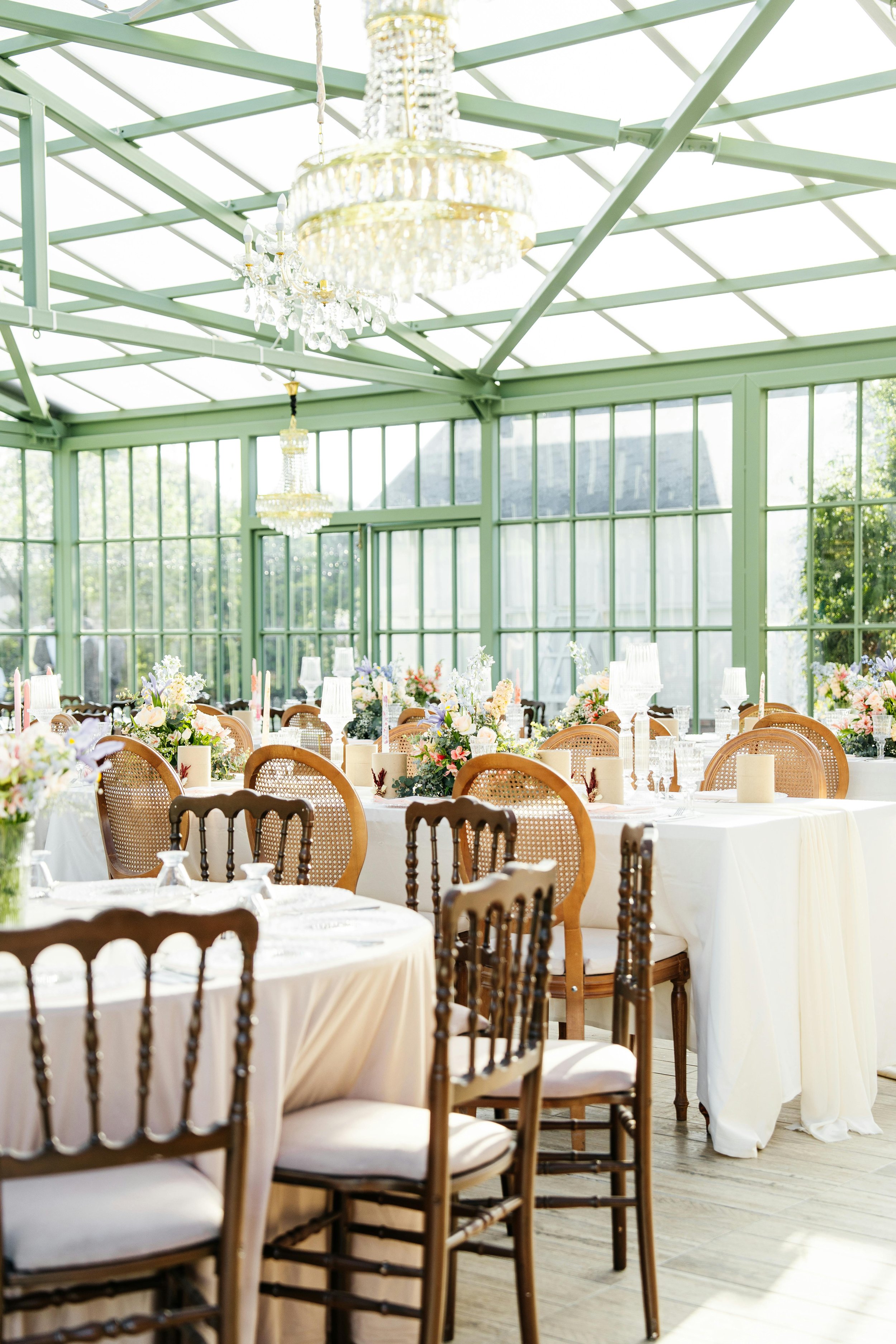 Elegant indoor event space with round tables covered in cream tablecloths, adorned with floral centerpieces, candles, and glassware. The room has large windows, a glass ceiling, and crystal chandeliers, creating a bright and airy atmosphere with wooden chairs around the tables.
