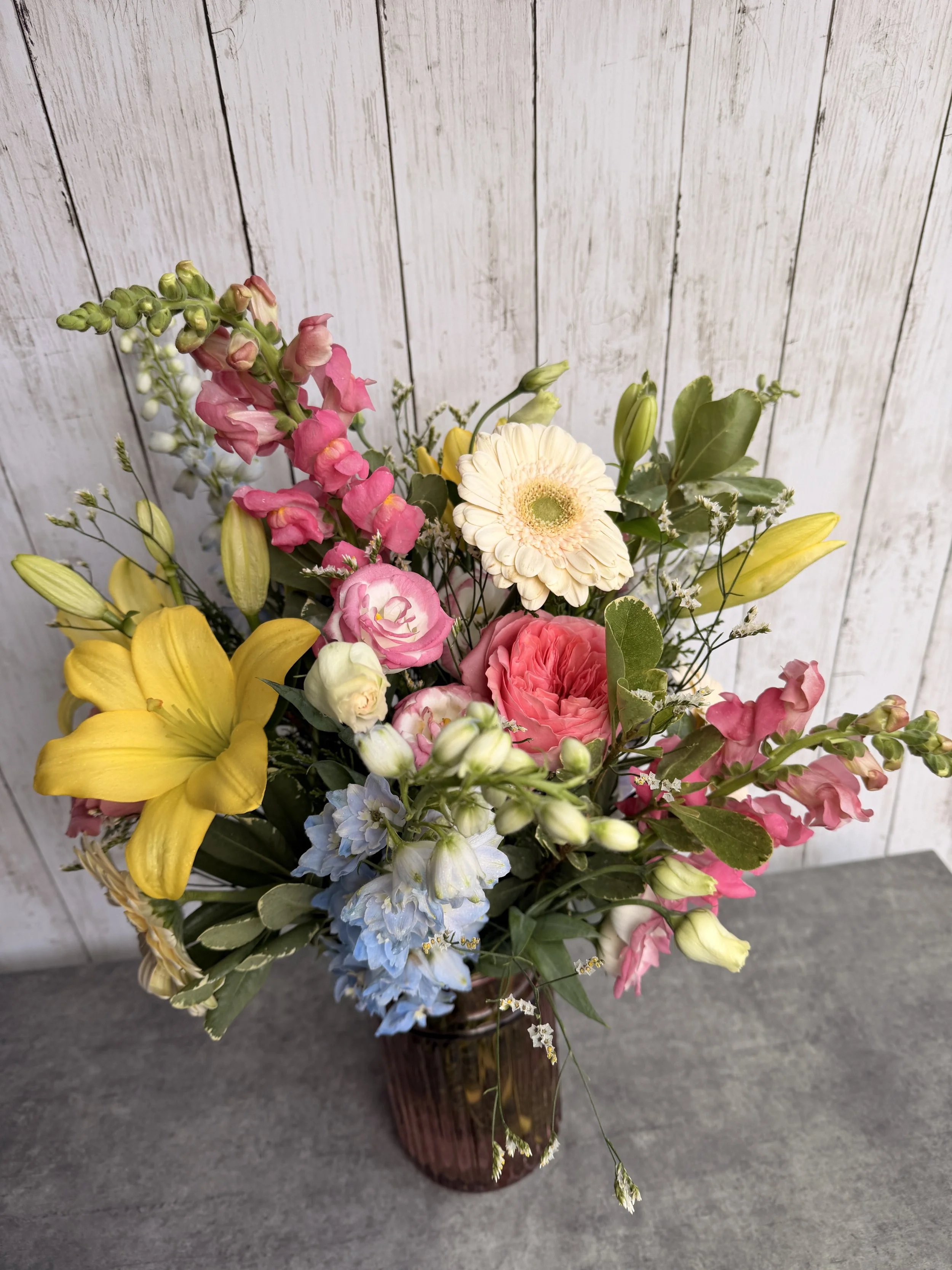 A colorful bouquet of various flowers in a brown vase on a gray surface against a white wooden panel background.