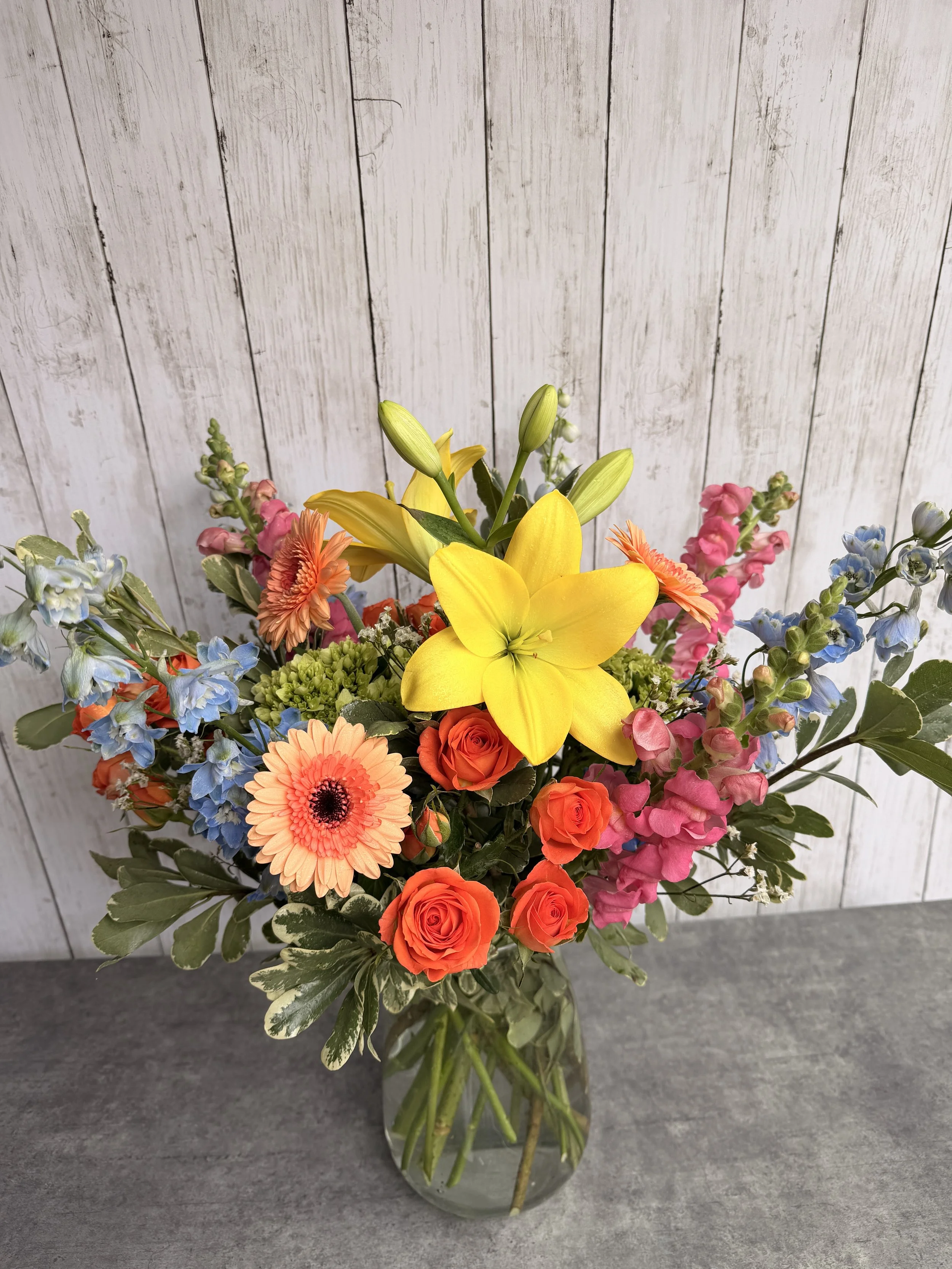 A colorful bouquet of flowers in a glass vase, featuring yellow lilies, peach gerbera daisies, orange roses, pink snapdragons, blue delphiniums, and other mixed greenery, set against a white wooden background.