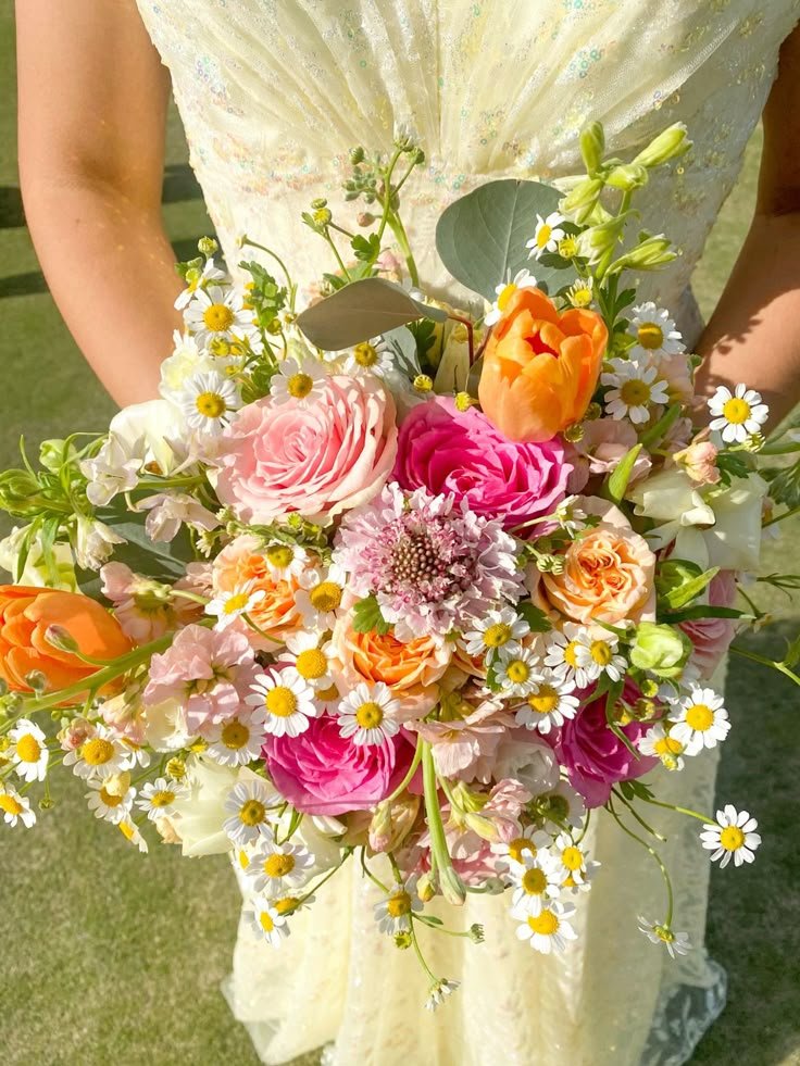 A person in a light yellow dress holding a bouquet of pink, orange, and white flowers with greenery, including daisies and roses.