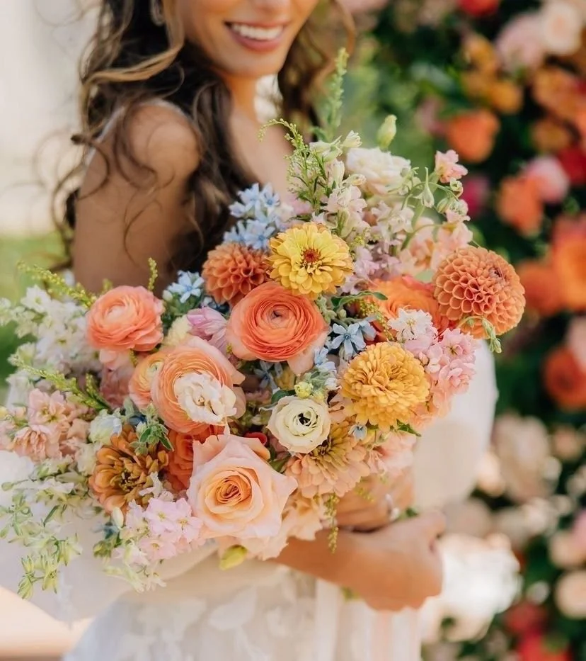 A woman holding a large bouquet of mixed colorful flowers, including roses, dahlias, and other blooms, outdoors with a blurred floral background.