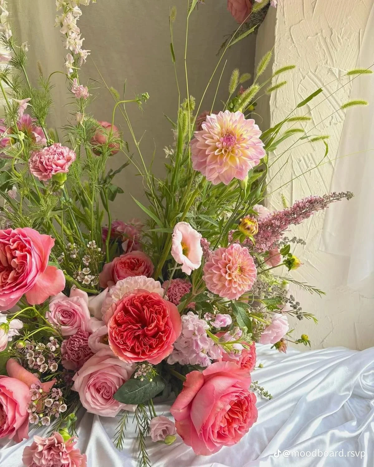 Decorative table setting with pink and peach flowers in glass vases, glasses of water, gold utensils, and a white plate with a large pink flower, on a pink napkin with a light blue tablecloth.