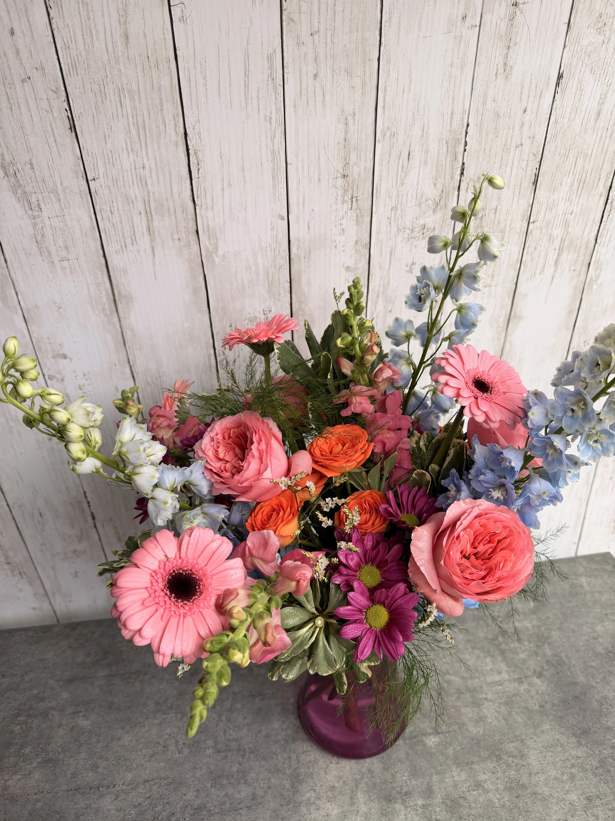 A colorful bouquet of flowers in a pink vase, featuring pink gerbera daisies, pink roses, orange ranunculus, purple daisies, blue delphiniums, and greenery, set against a white wooden background.