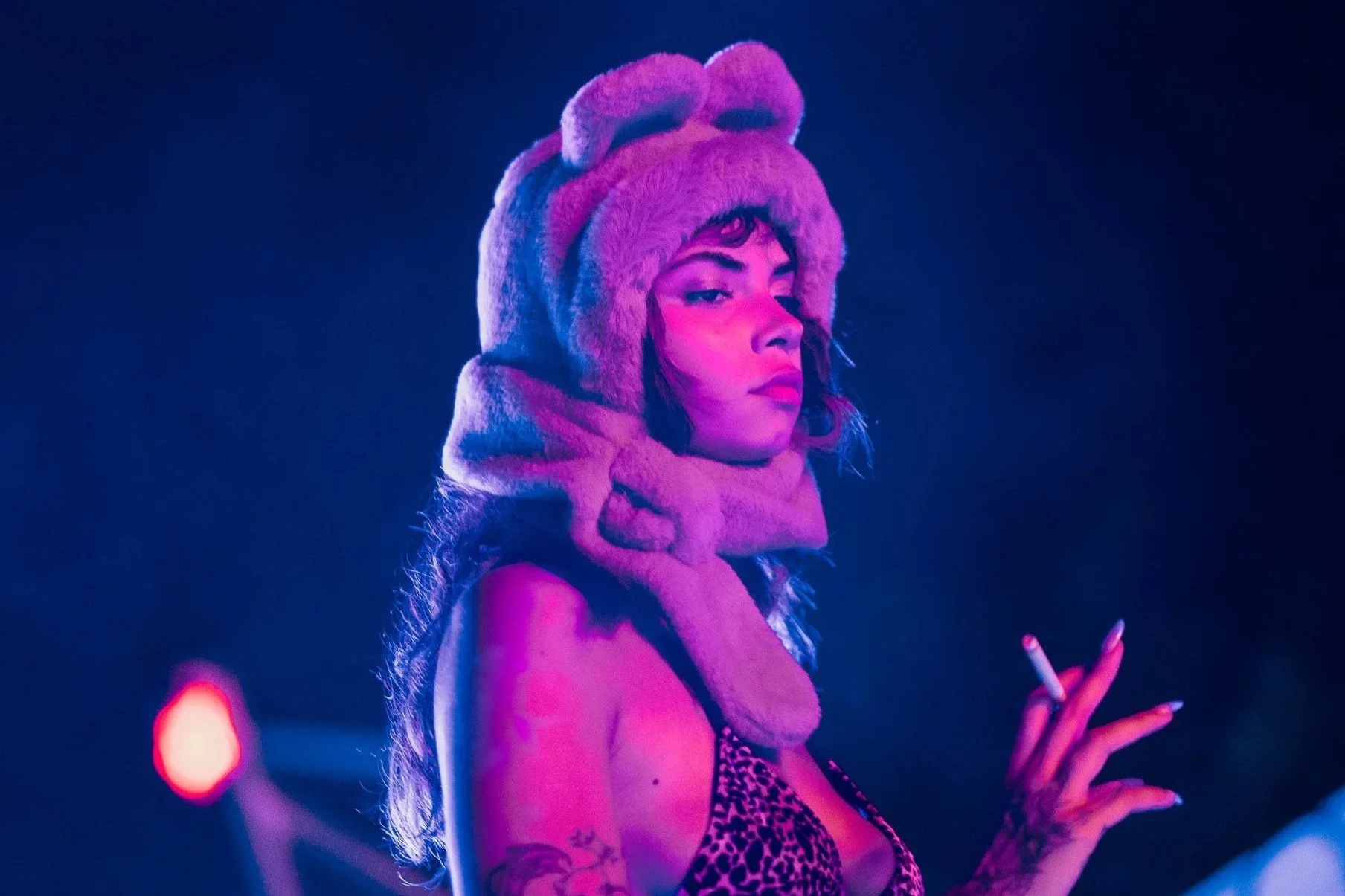 A woman wearing a pink furry animal hat and a leopard print top, holding a cigarette, with colorful stage lighting in the background.