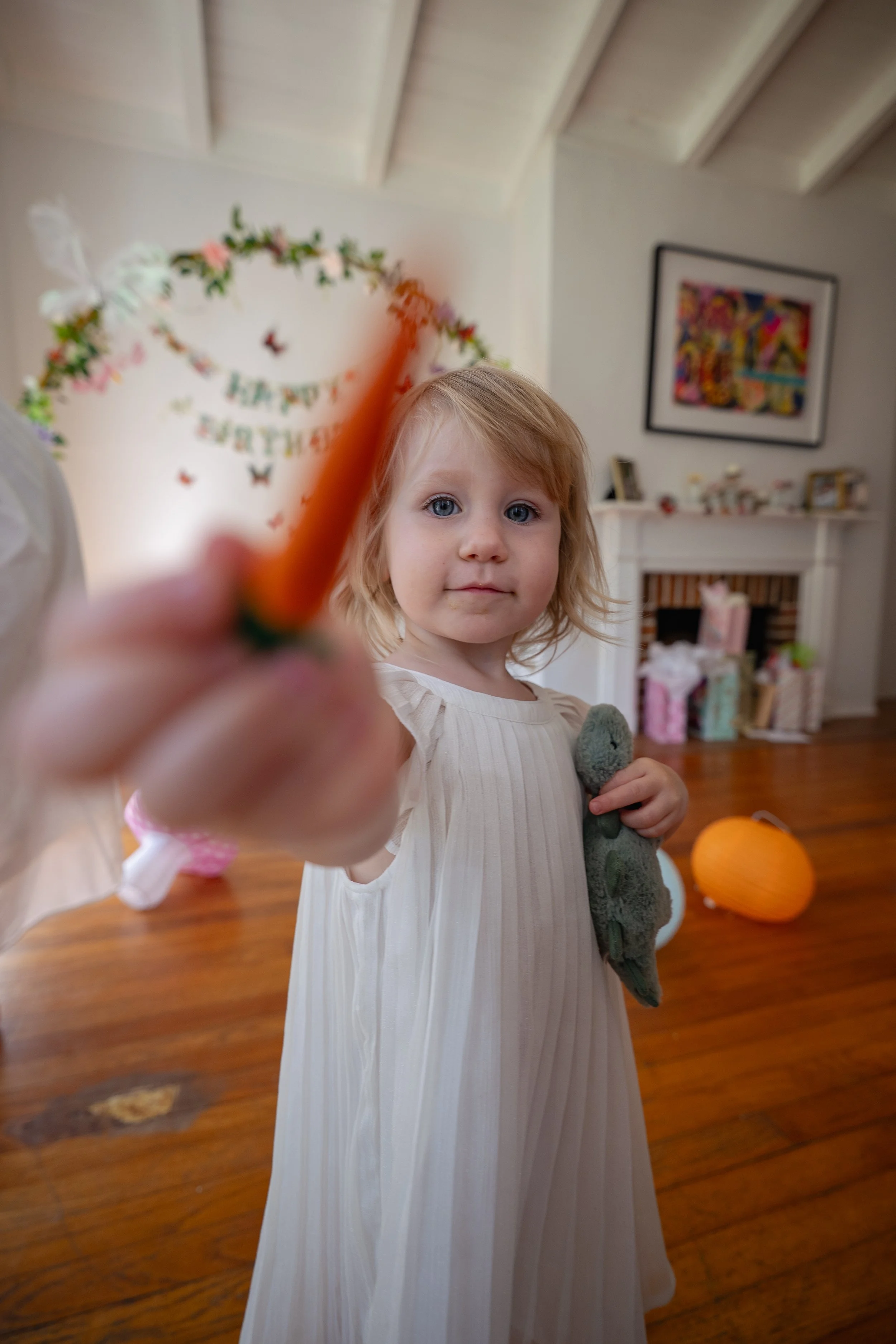 A young girl with blonde hair and blue eyes, wearing a white dress, is holding a stuffed animal in one hand and an orange object, possibly a carrot, towards the camera with the other hand inside a decorated room celebrating a birthday.