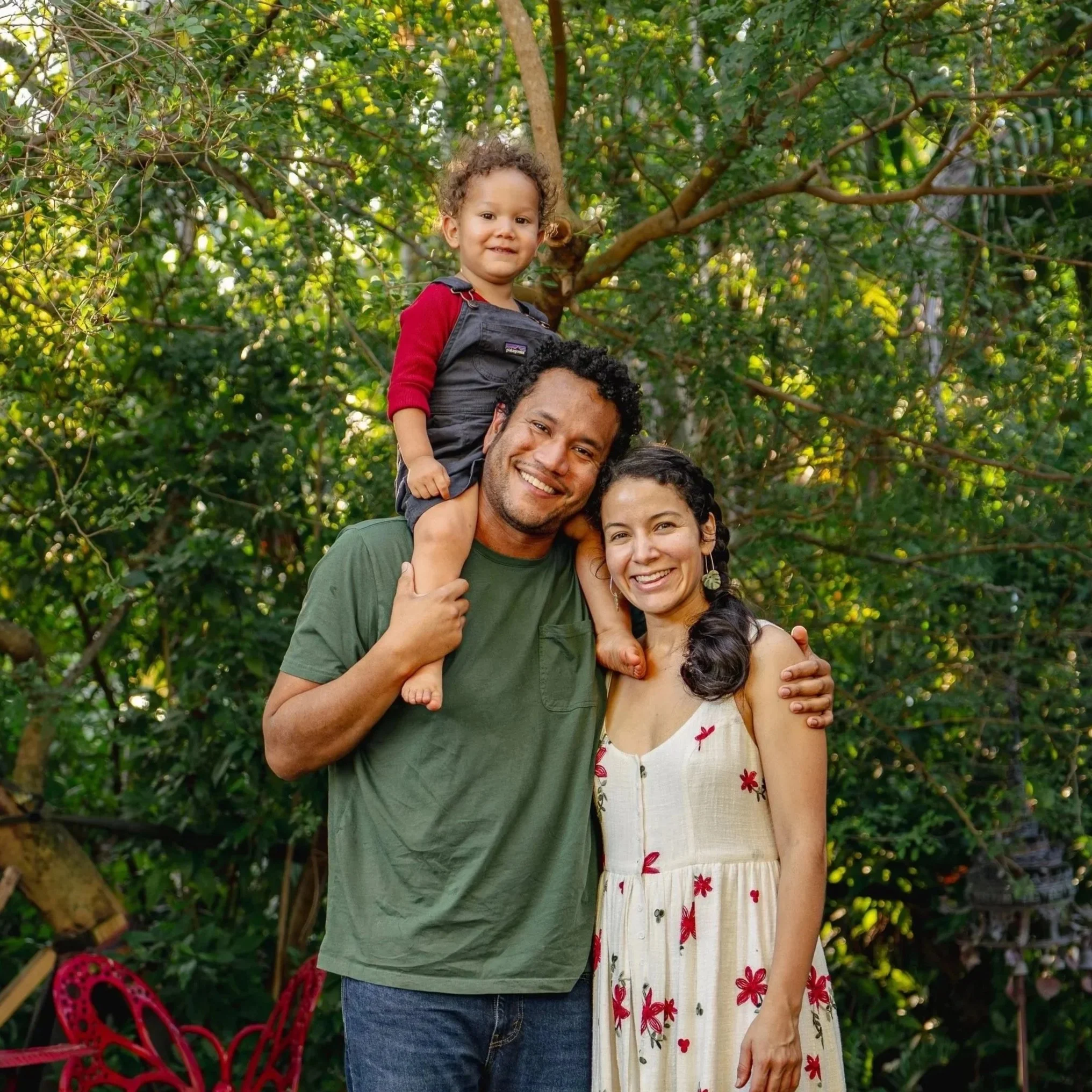 A happy family of three outdoors in a lush green garden, with a man carrying a young girl on his shoulders and a woman standing next to them, all smiling.