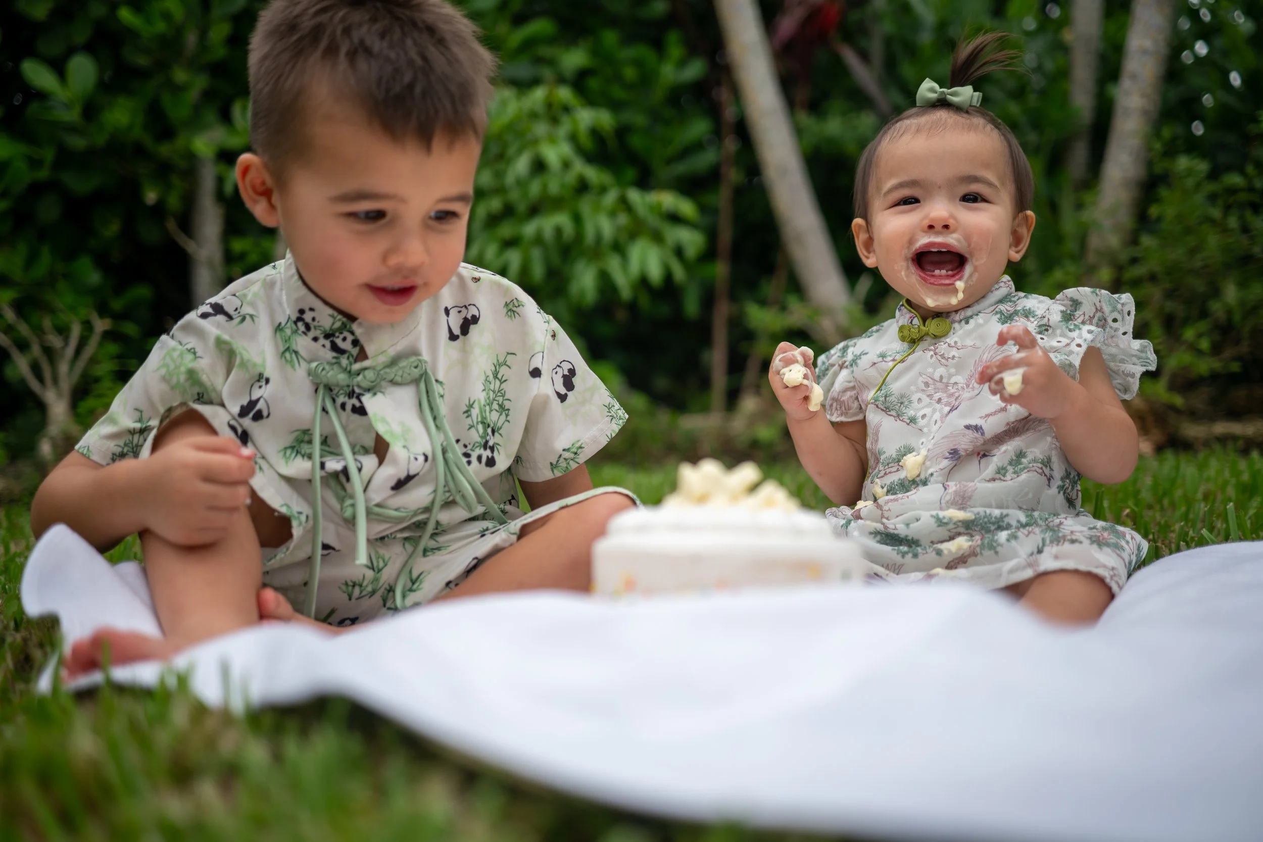 Two young children celebrating a birthday outdoors, sitting on a white blanket with a cake in front of them, surrounded by greenery.