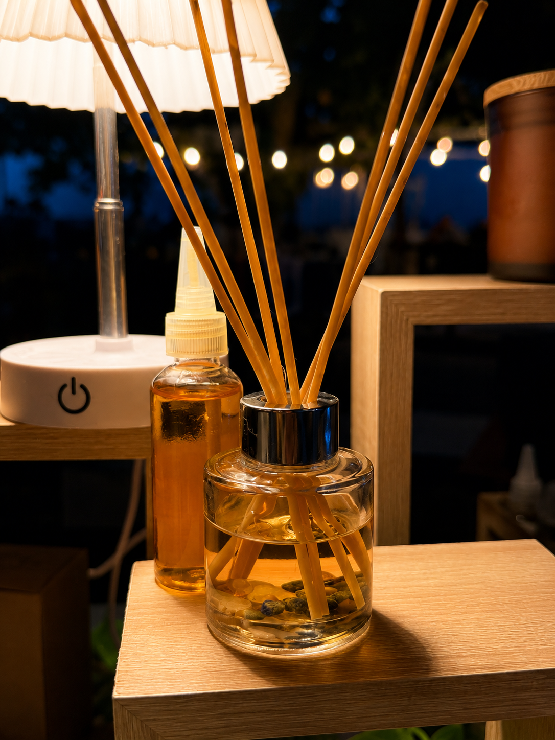 Vela encendida con tono cálido, frascos con aceite y difusor de varillas en un ambiente de noche o interior con luces en el fondo.