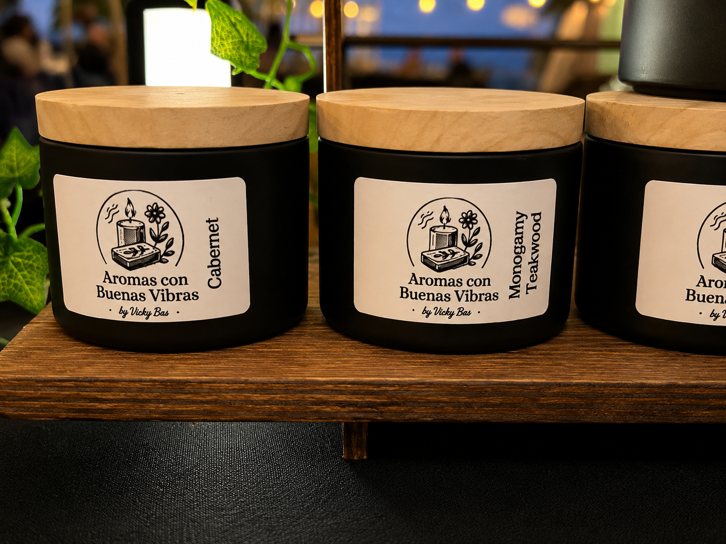 Tres frascos negros con tapas de madera, cada uno con una etiqueta que dice aromas con buenas vibras, uno con texto cabernet y otro con texto monogamy teakwood, sobre una superficie de madera.