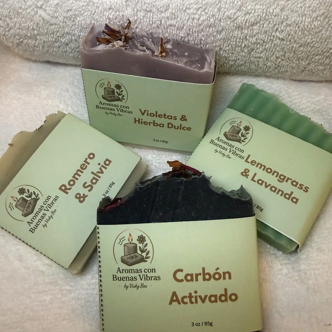 Cuatro barras de jabón con diferentes aromas, uno de ellos con carbón activado, las otras con violetas y hierba dulce, romero y salvia, y lemongrass y lavanda, colocados sobre una toalla beige.