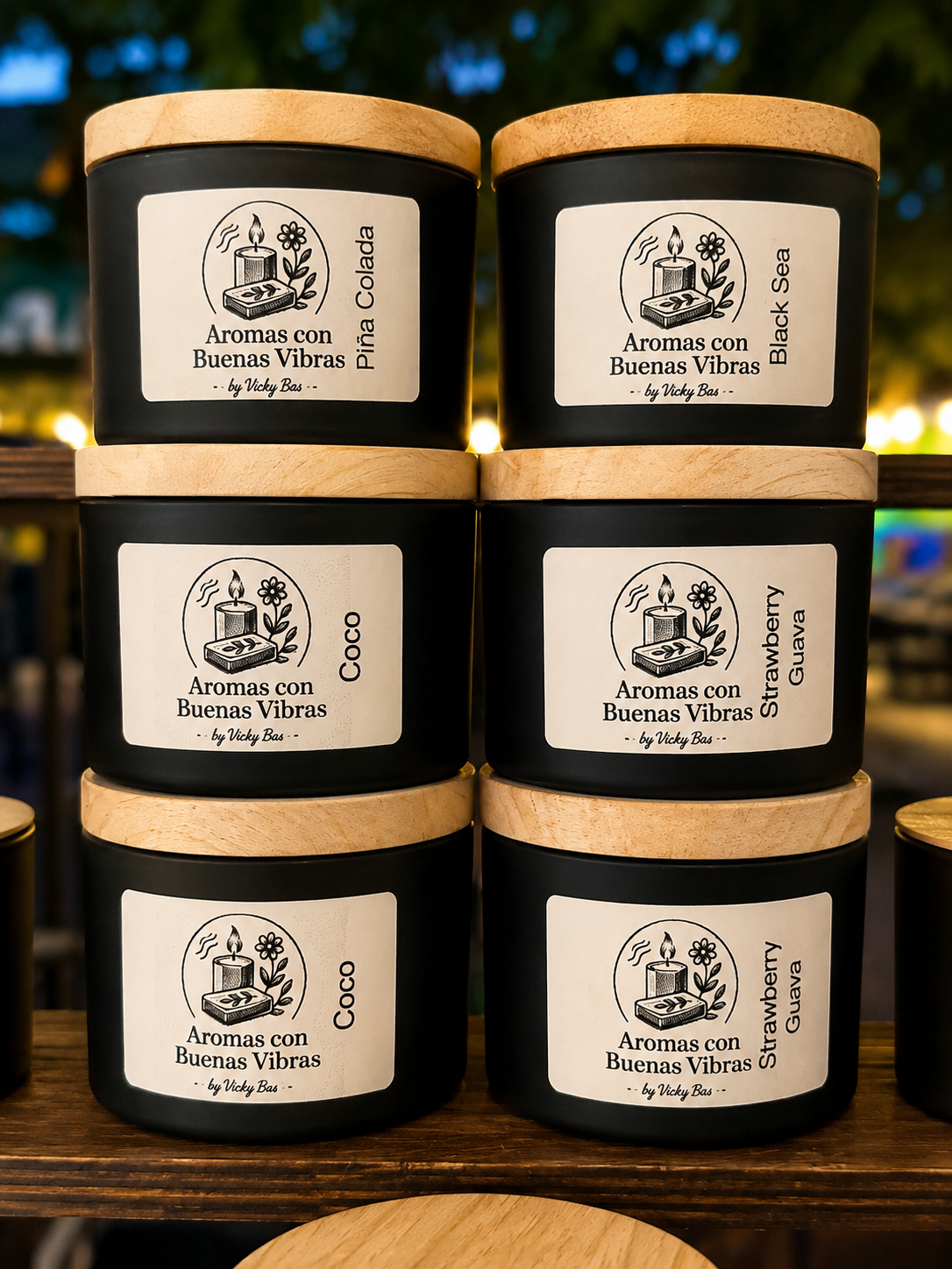 Latas negras con tapas de madera, etiquetadas con diferentes aromas: Piña Collada, Black Sea, Coco y Strawberry Guava, con un dibujo de vela y flores en la etiqueta.