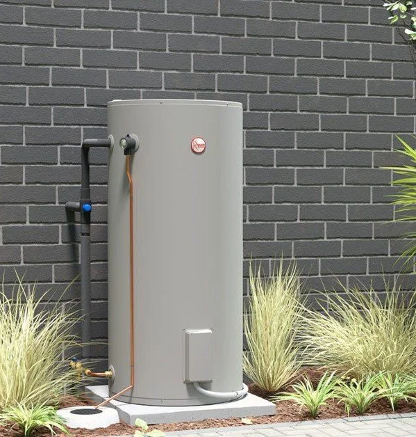 A large outdoor grey water heater with pipes connected to it, set against a dark brick wall with some ornamental grass plants nearby.