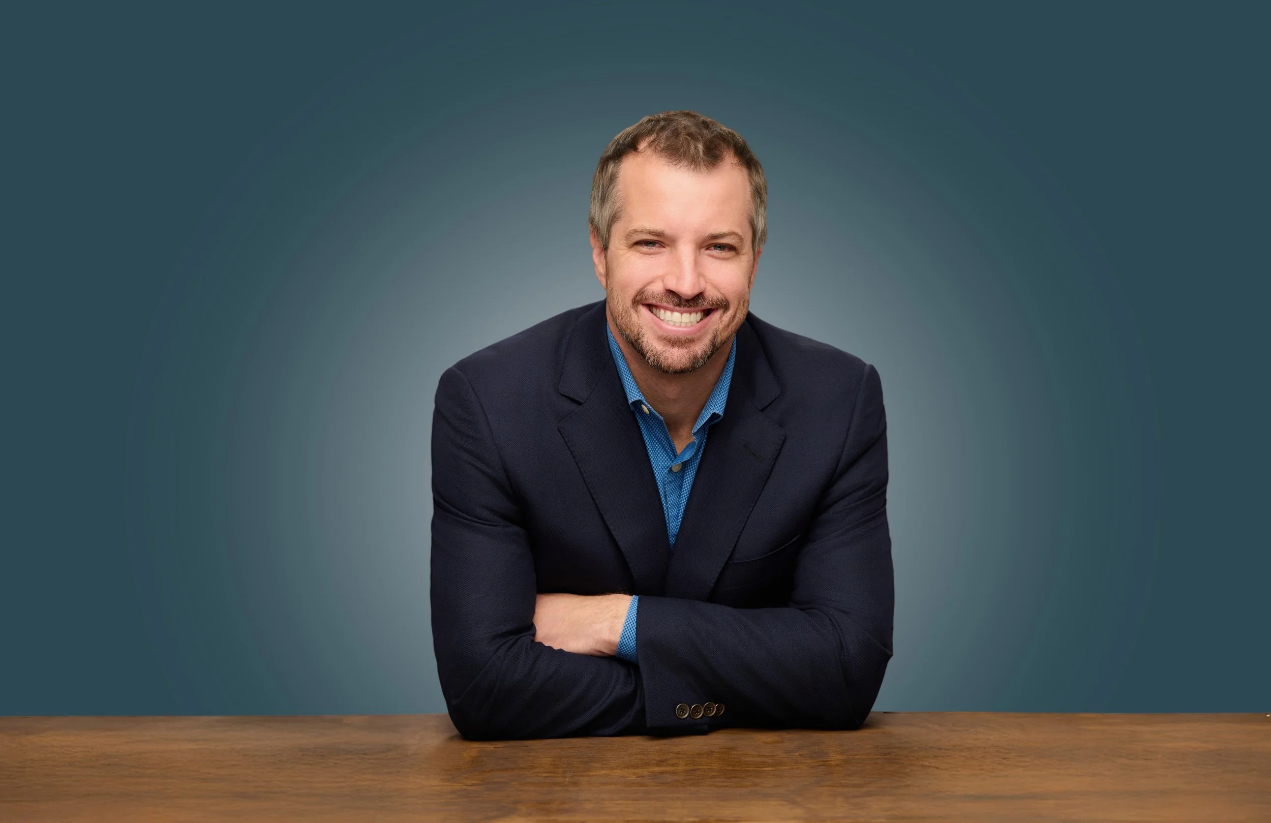 A smiling man with a short beard and mustache, wearing a dark blazer and blue shirt, sitting at a wooden table against a gradient blue background.