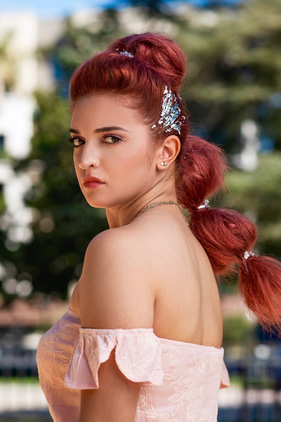 A photoshoot of a young woman with red hair styled in a curly ponytail, adorned with silver decorations, wearing pearl earrings, and an off-the-shoulder pink dress, outdoors with blurred trees and buildings in the background.
